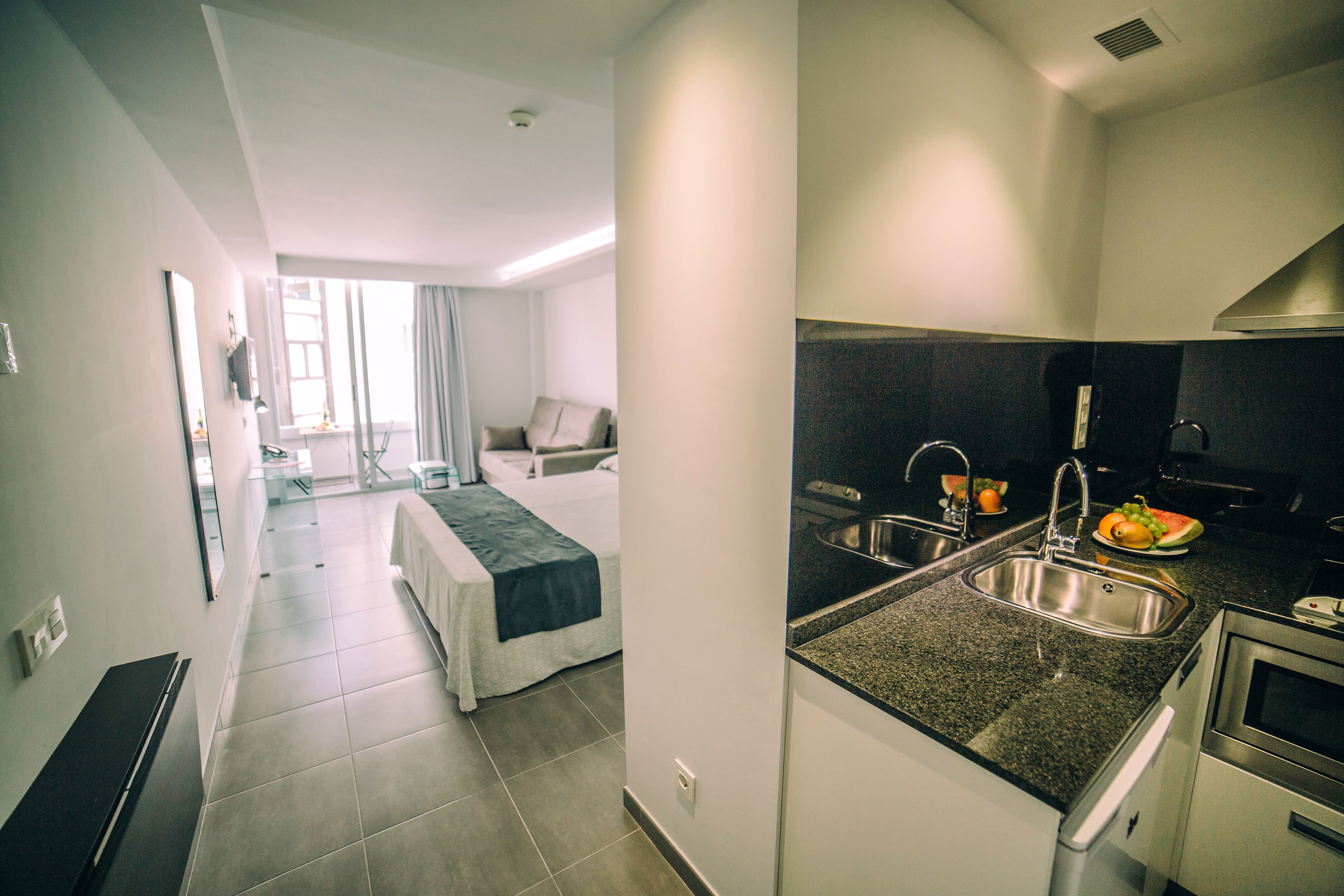 Quarto de hotel moderno com cozinha compacta, cama com colcha cinza e branca, um pequeno espaço de estar com poltrona e mesa, e vista para o corredor e outro quarto.