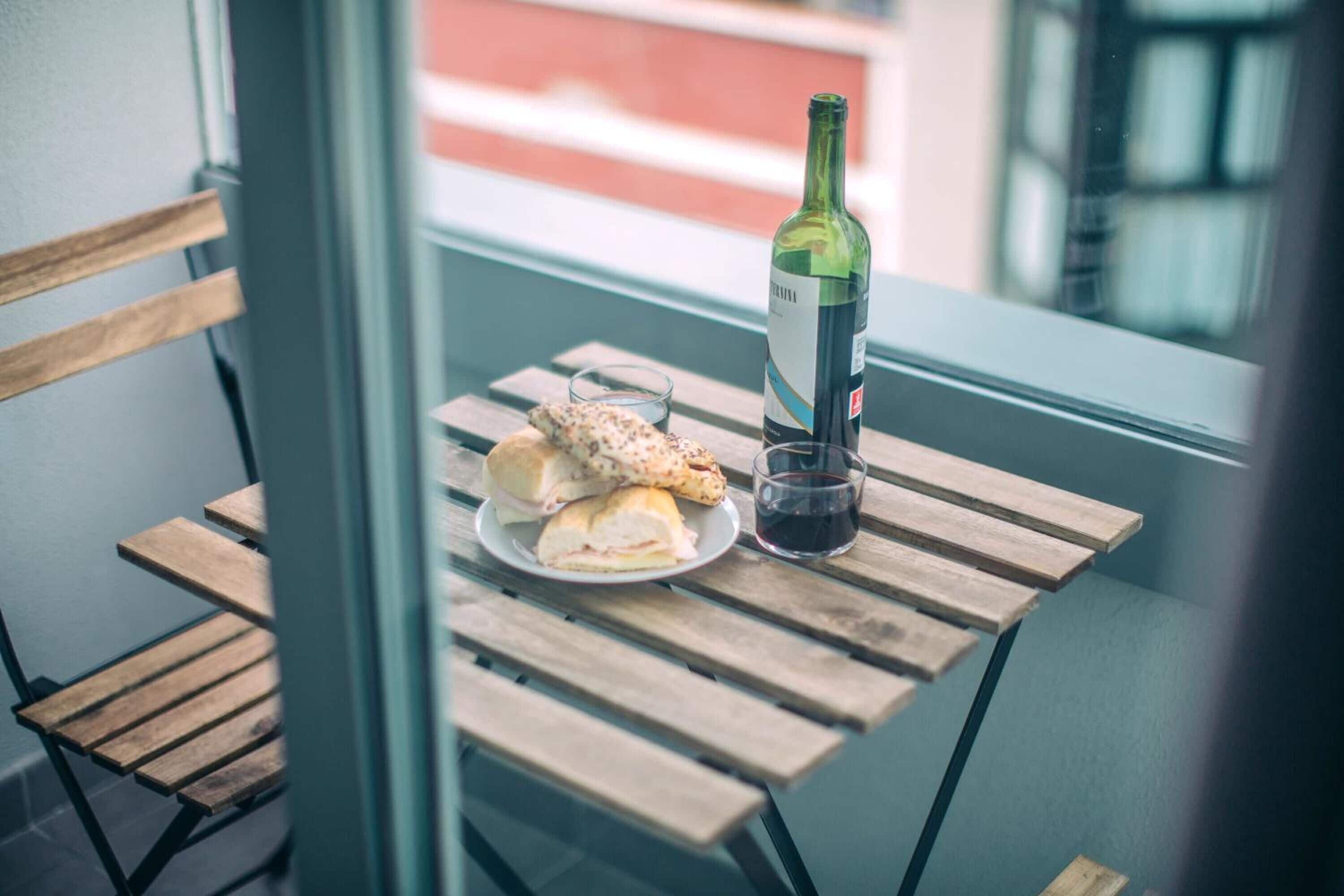 A wooden table with a bottle of wine and a plate of sandwiches on a balcony.