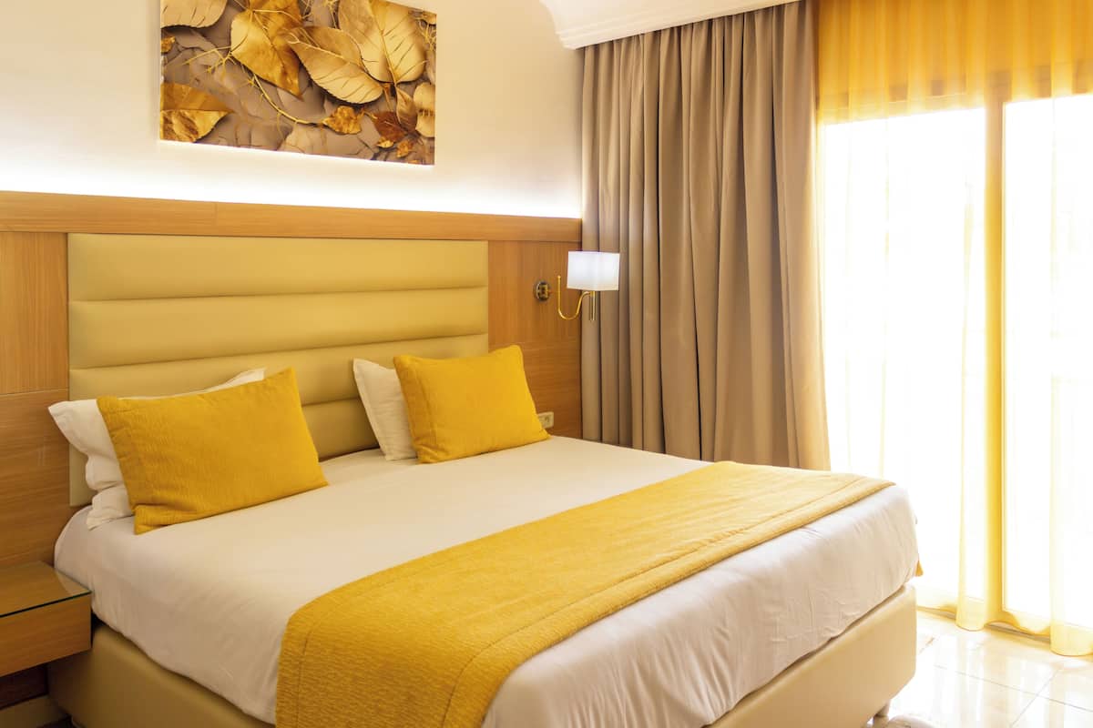 A hotel room with a large bed, a nightstand, a wall-mounted lamp, and a window with curtains.