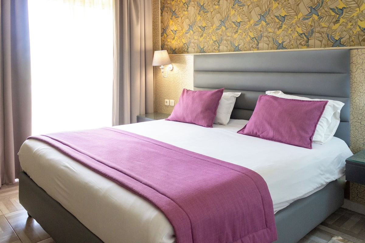 A hotel room with a large bed, a nightstand with a lamp, and a patterned wallpaper.
