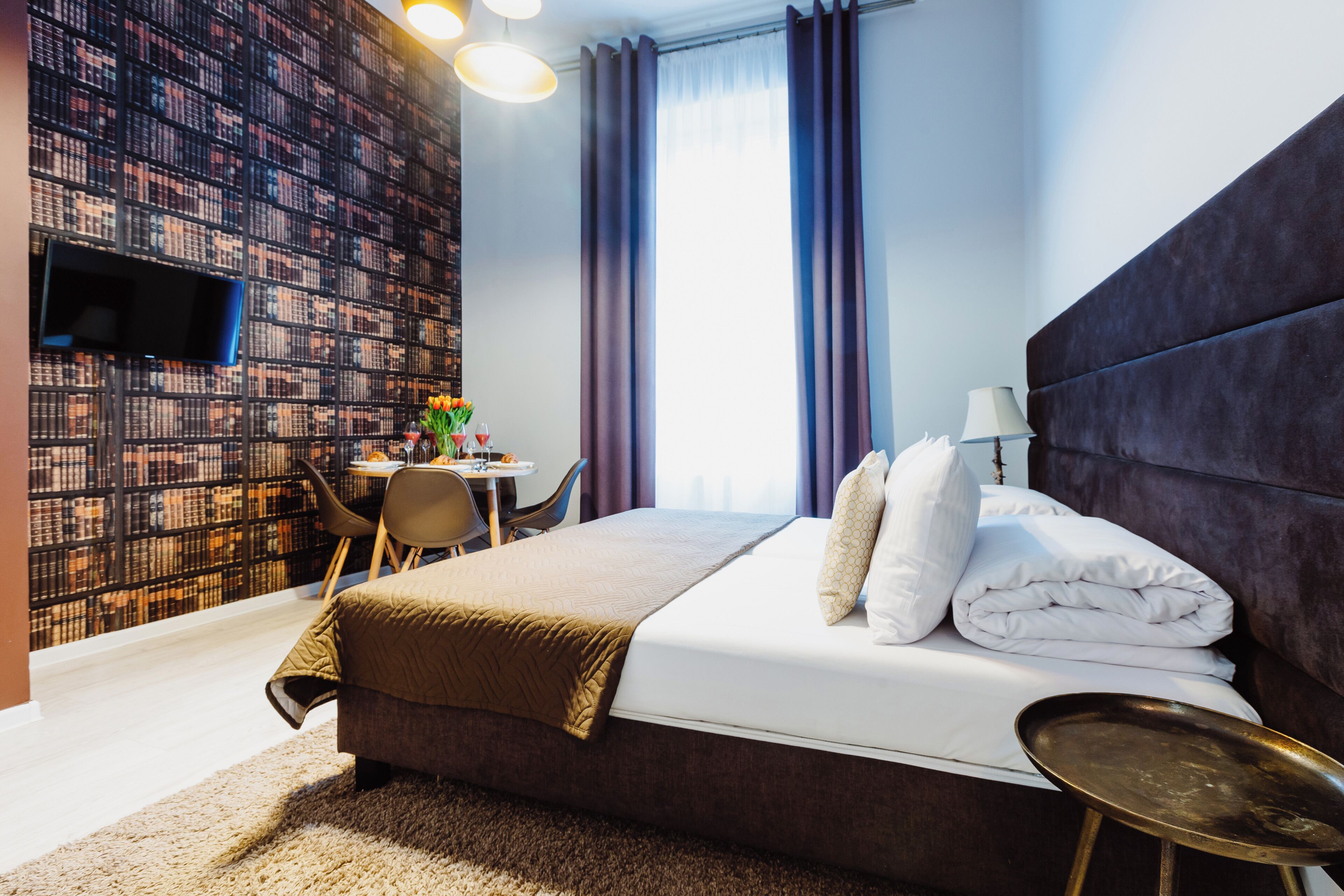 Ein Hotelzimmer mit einem großen Bett, einem Schreibtisch mit Stuhl, einem Bücherregal und einem an der Wand befestigten Fernseher.