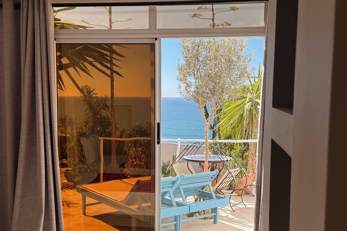 Un dormitorio con cama, una colcha roja y vistas a un balcón exterior con una mesa y sillas.
