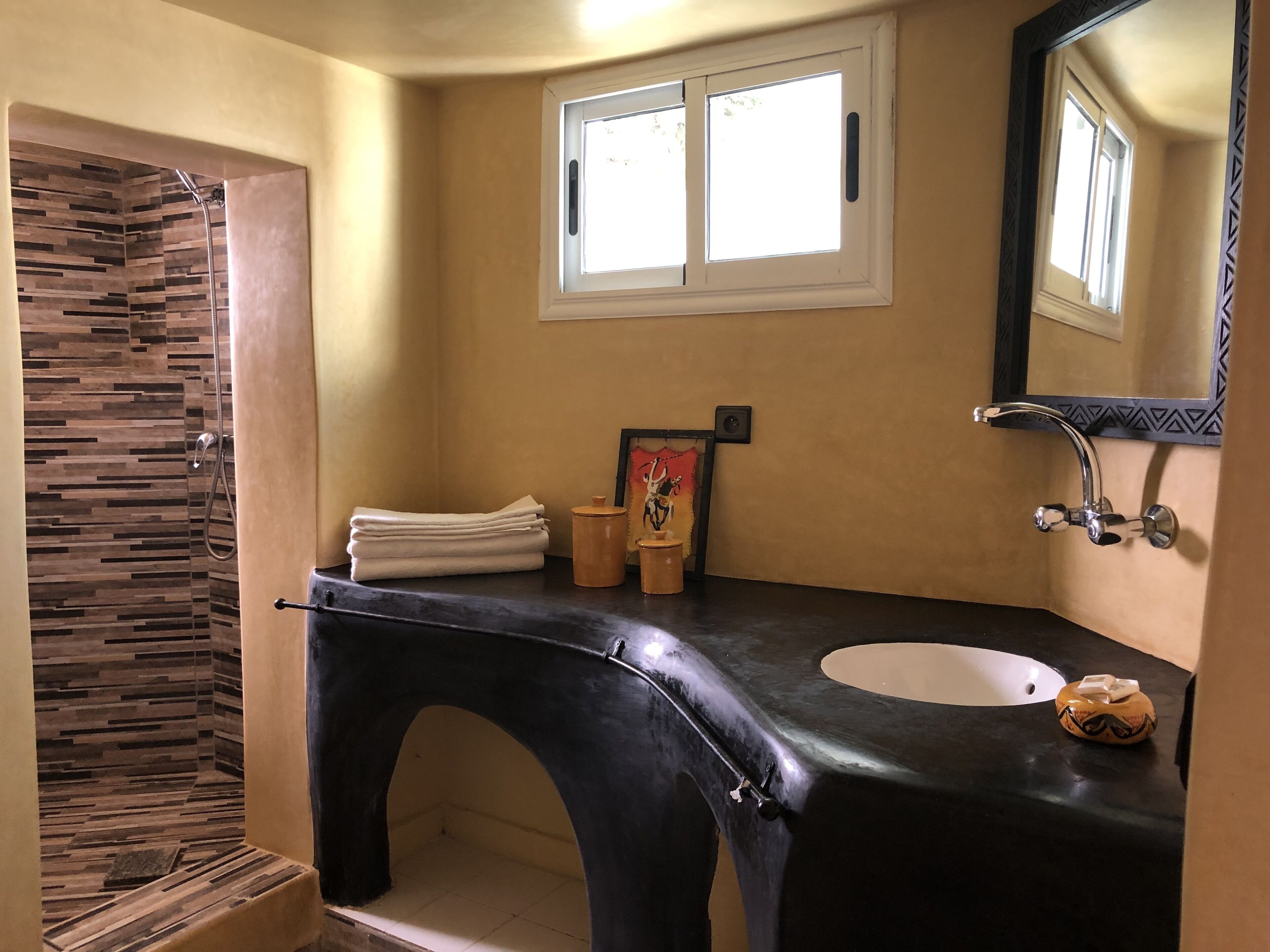 A bathroom with a black countertop sink, a shower with a tiled wall, and a window above the mirror.
