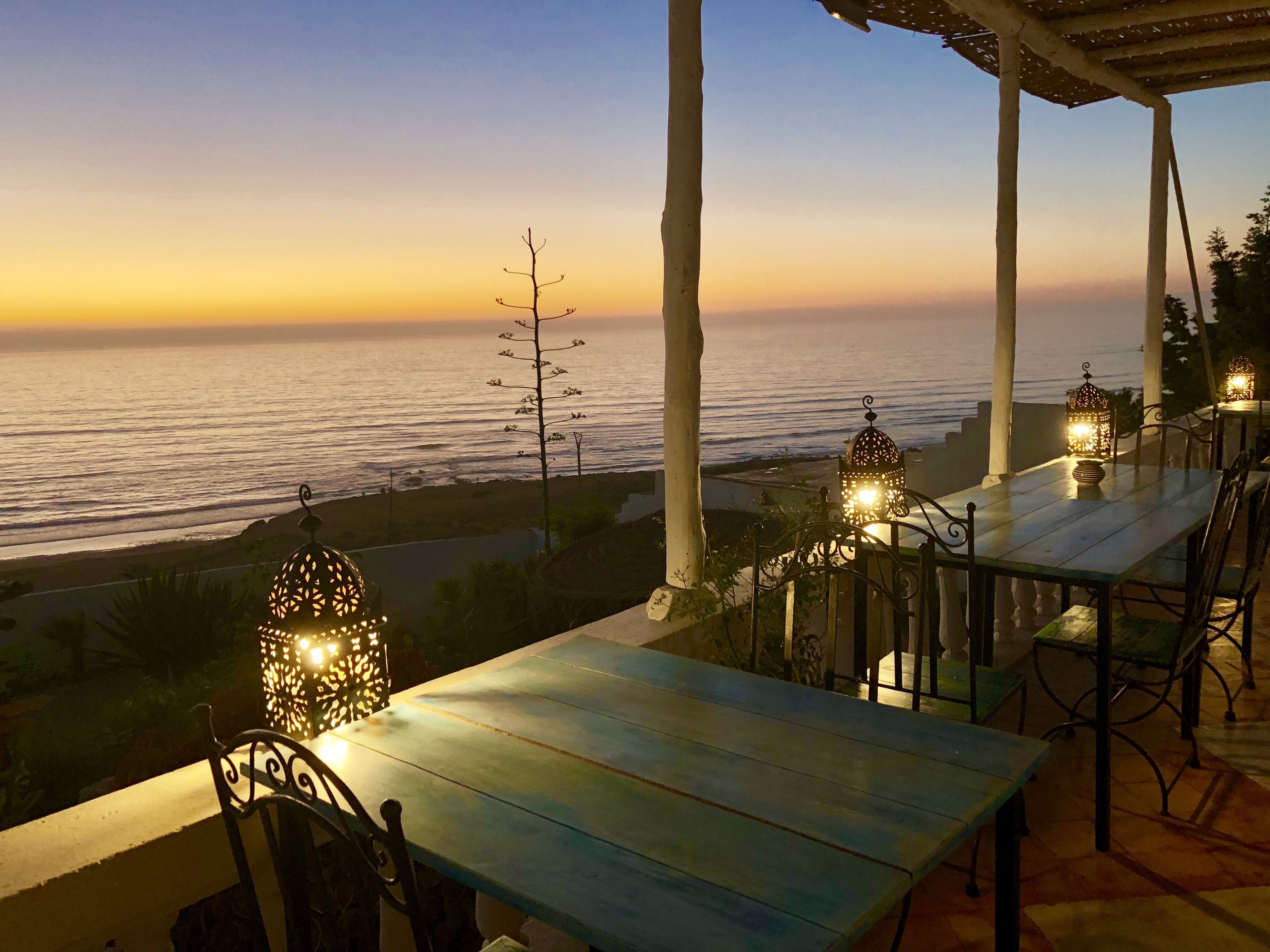 An outdoor seating area with tables and chairs, overlooking a beach at sunset.