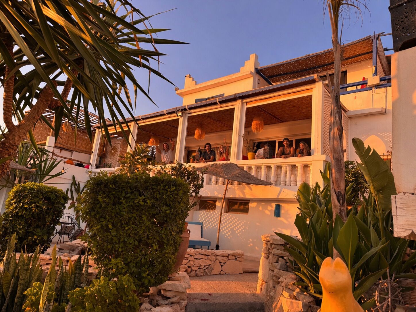 A two-story building with a balcony, surrounded by tropical plants and a stone wall.
