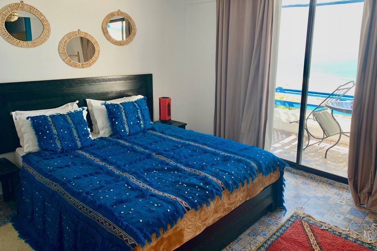 Un dormitorio con cama, mesita de noche, lámpara roja y vistas a un espacio exterior con una silla y una mesa.