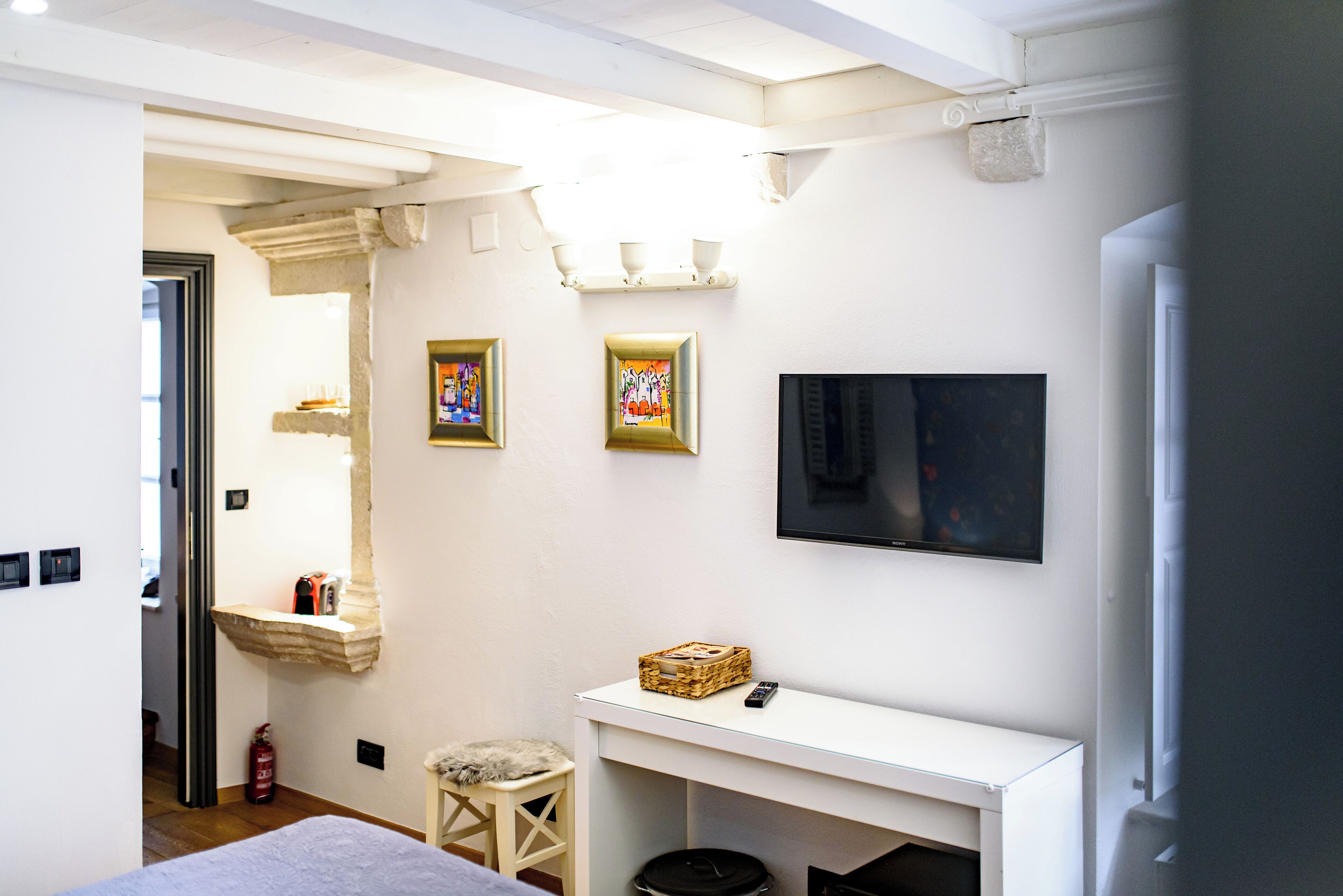 A hotel room with a white console table, a television mounted on the wall, and two framed pictures.