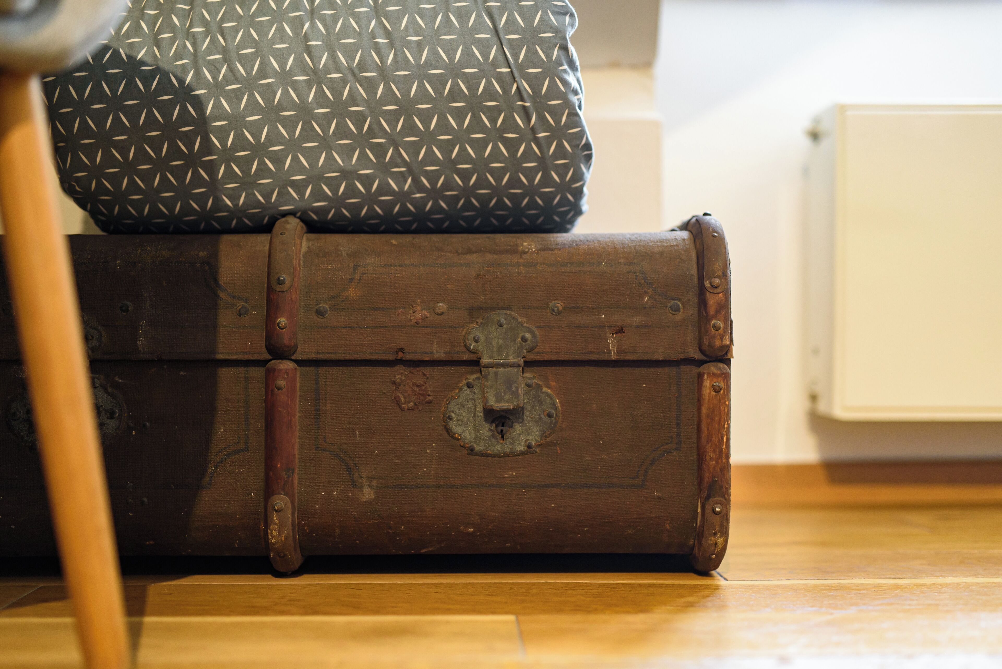 A wooden chest with a metal latch, a patterned fabric bag, and a wooden chair leg.