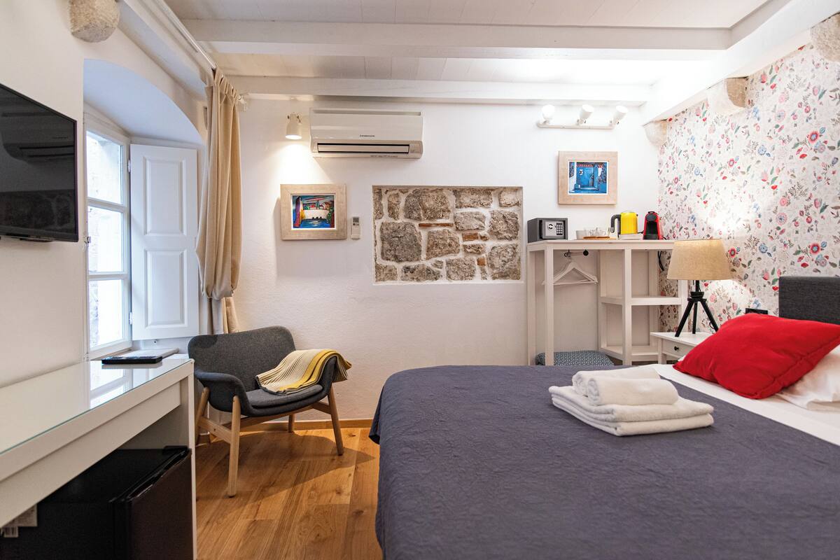 A bedroom with a bed, a chair, a desk, an air conditioner, and a wall-mounted TV.