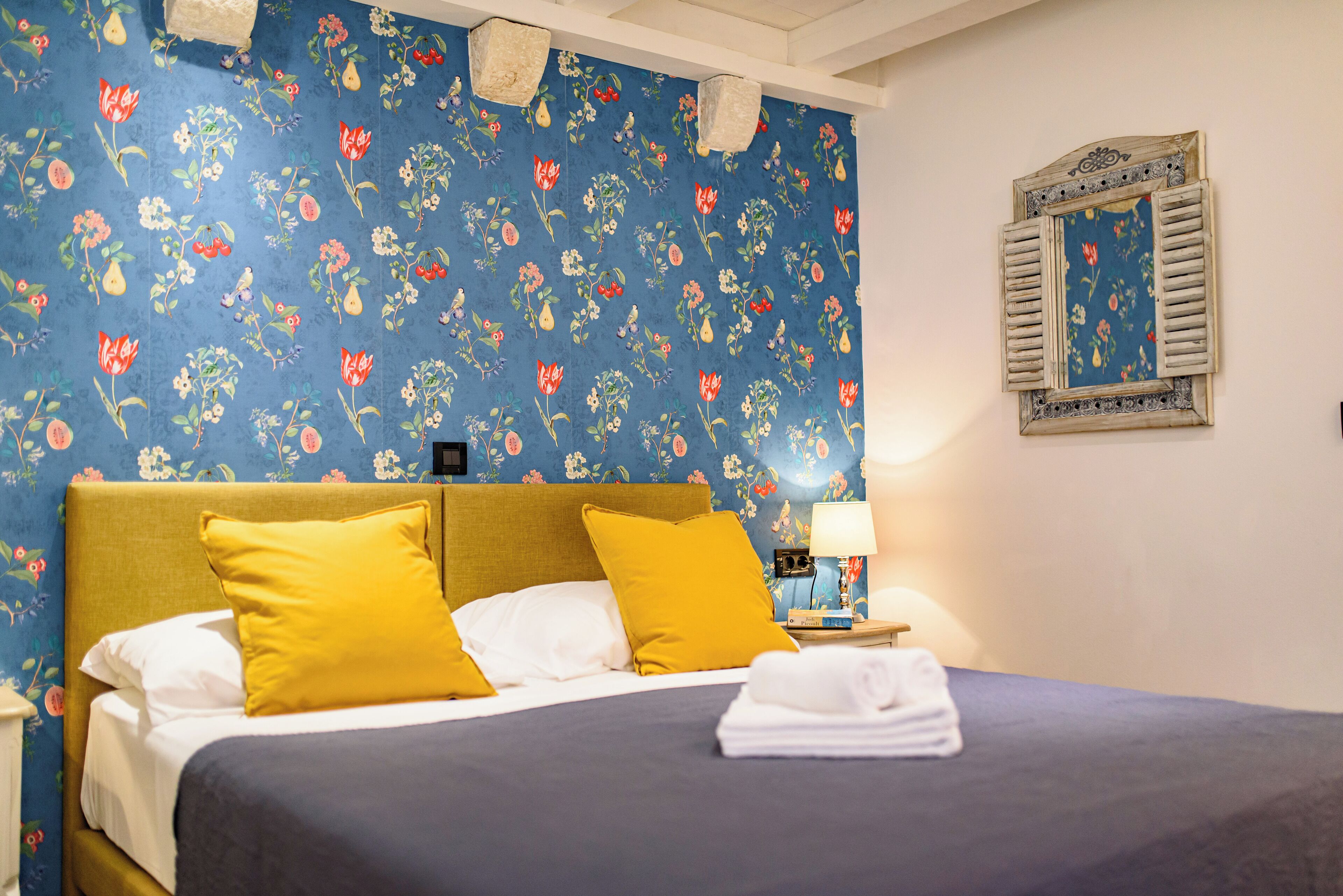 Uma cama bem arrumada com almofadas amarelas, papel de parede floral azul e um espelho emoldurado na parede.