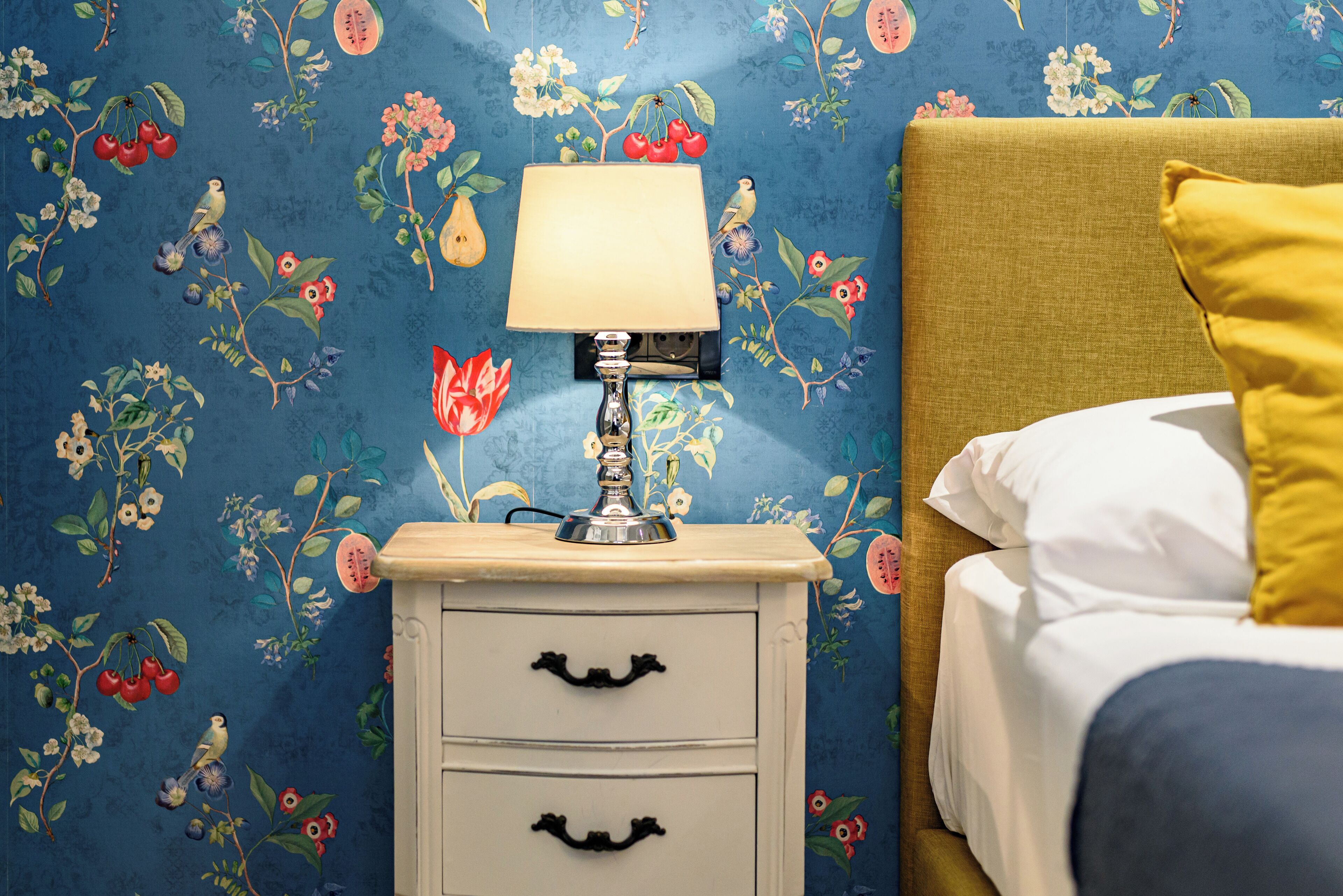 Un dormitorio con papel tapiz floral, una mesita de noche con lámpara y una cama con ropa de cama blanca y amarilla.