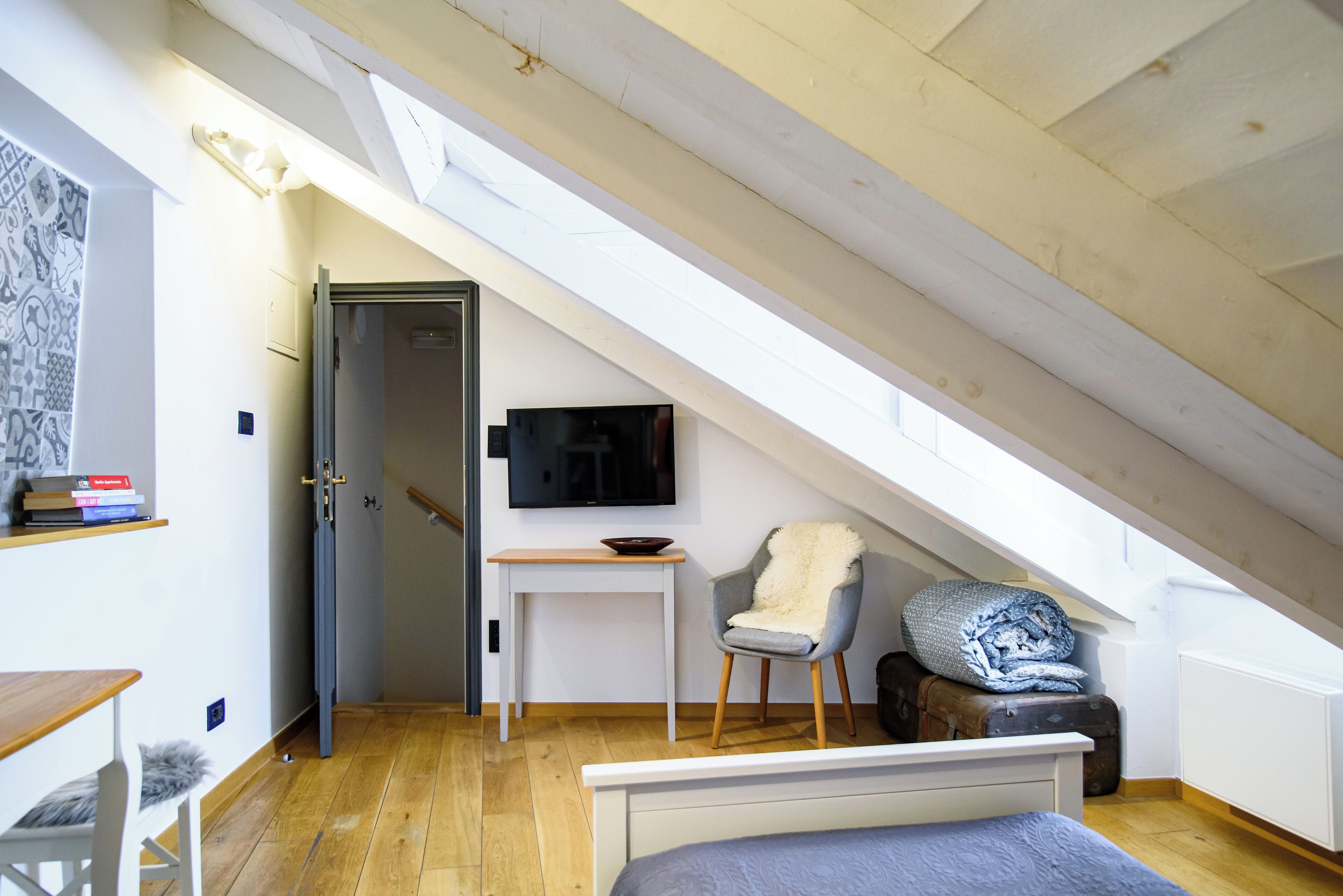 A small, well-lit attic bedroom with a sloped ceiling, a single bed, a desk, a chair, and a television.