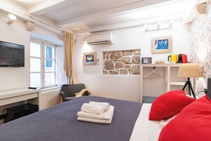 Quarto de hotel com cama, escrivaninha, cadeira, televisão, ar condicionado, parede de pedra e janela com cortinas.