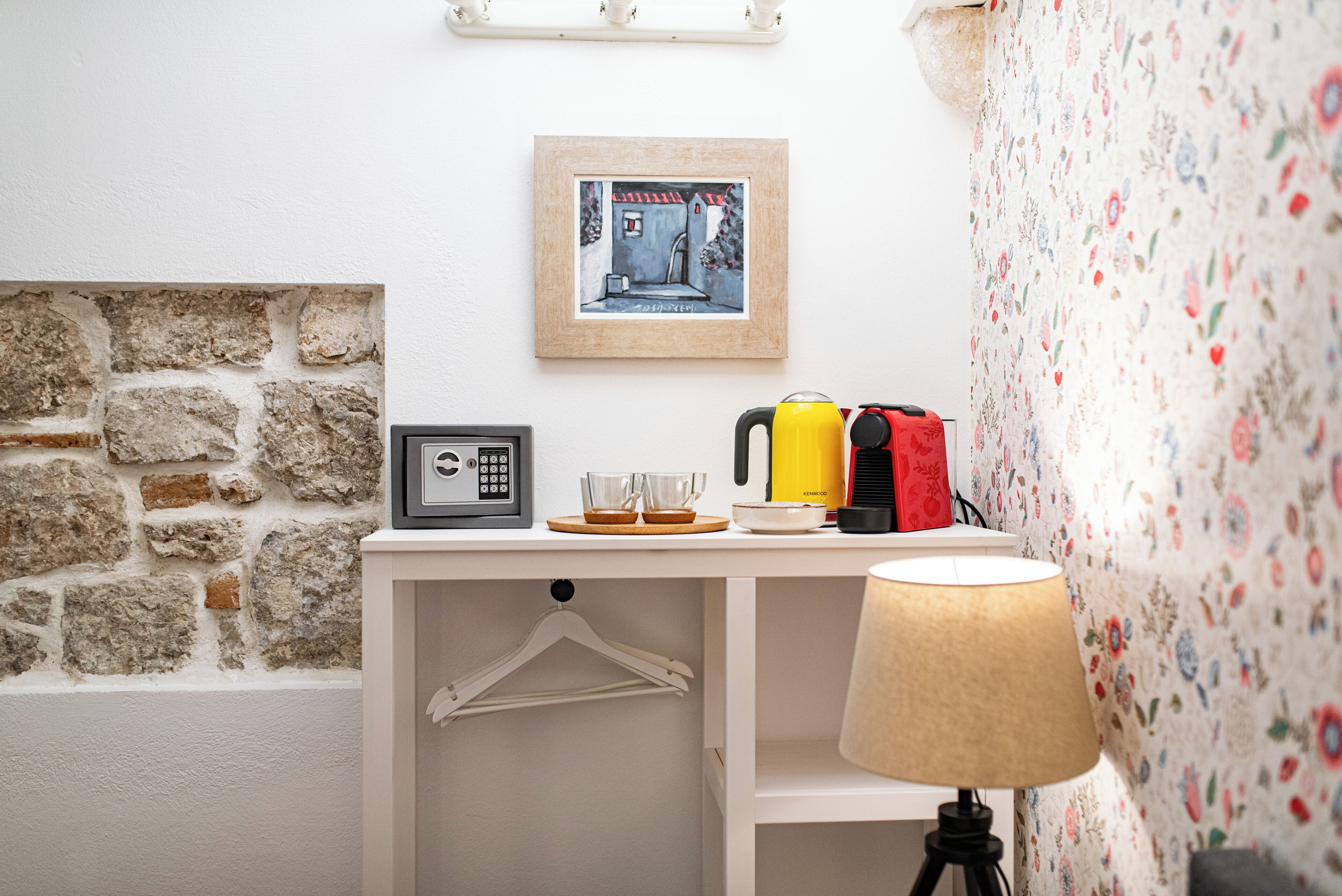 A small, neatly organized room with a stone wall, a framed picture, a kettle, a lamp, and a wall-mounted shelf.