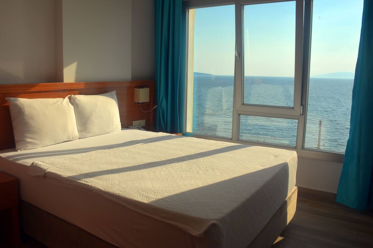 A hotel room with a large bed, a nightstand, a window with a view of the sea, and a wooden headboard.