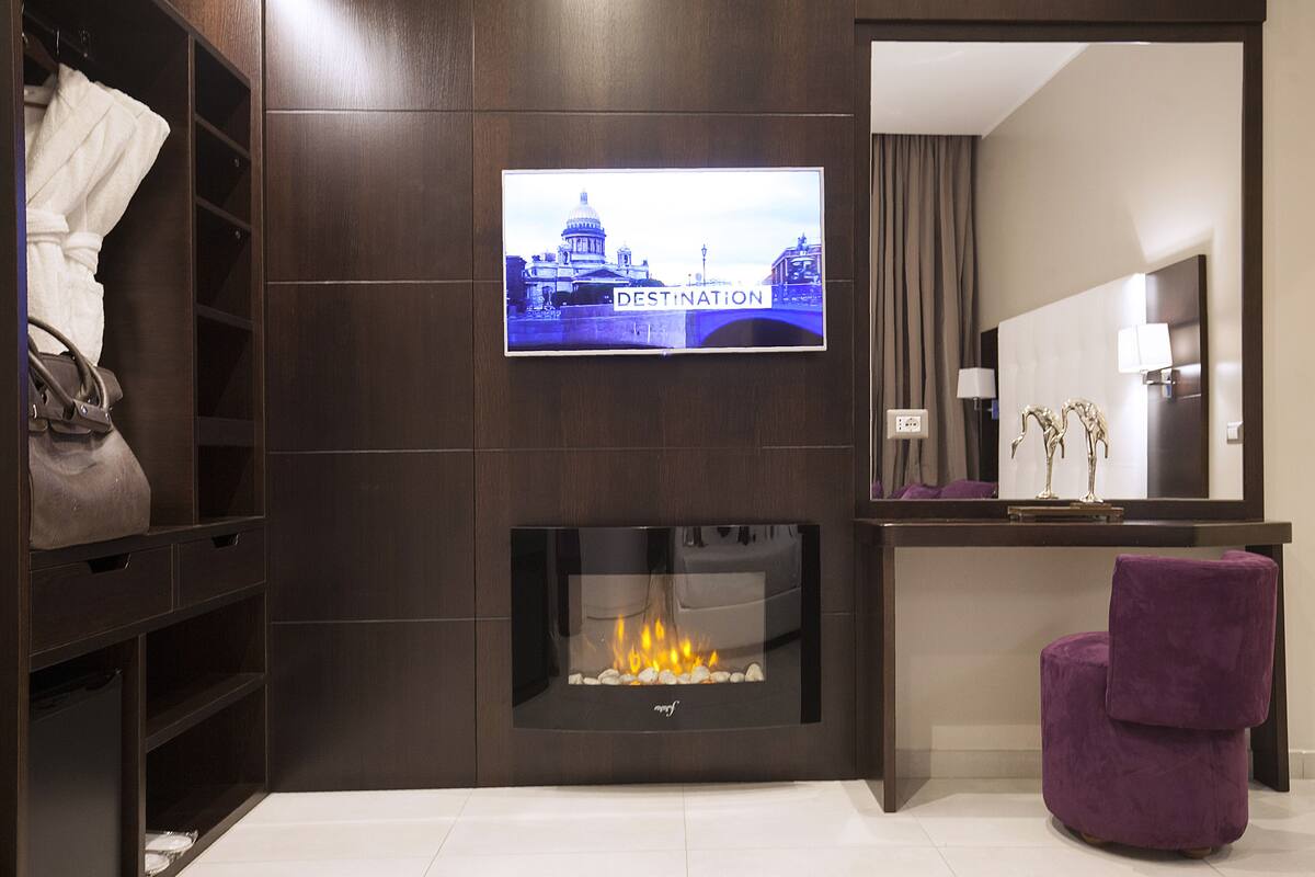 Habitación de hotel moderna con una televisión que muestra un paisaje urbano, una chimenea, un tocador con espejo y una silla morada.