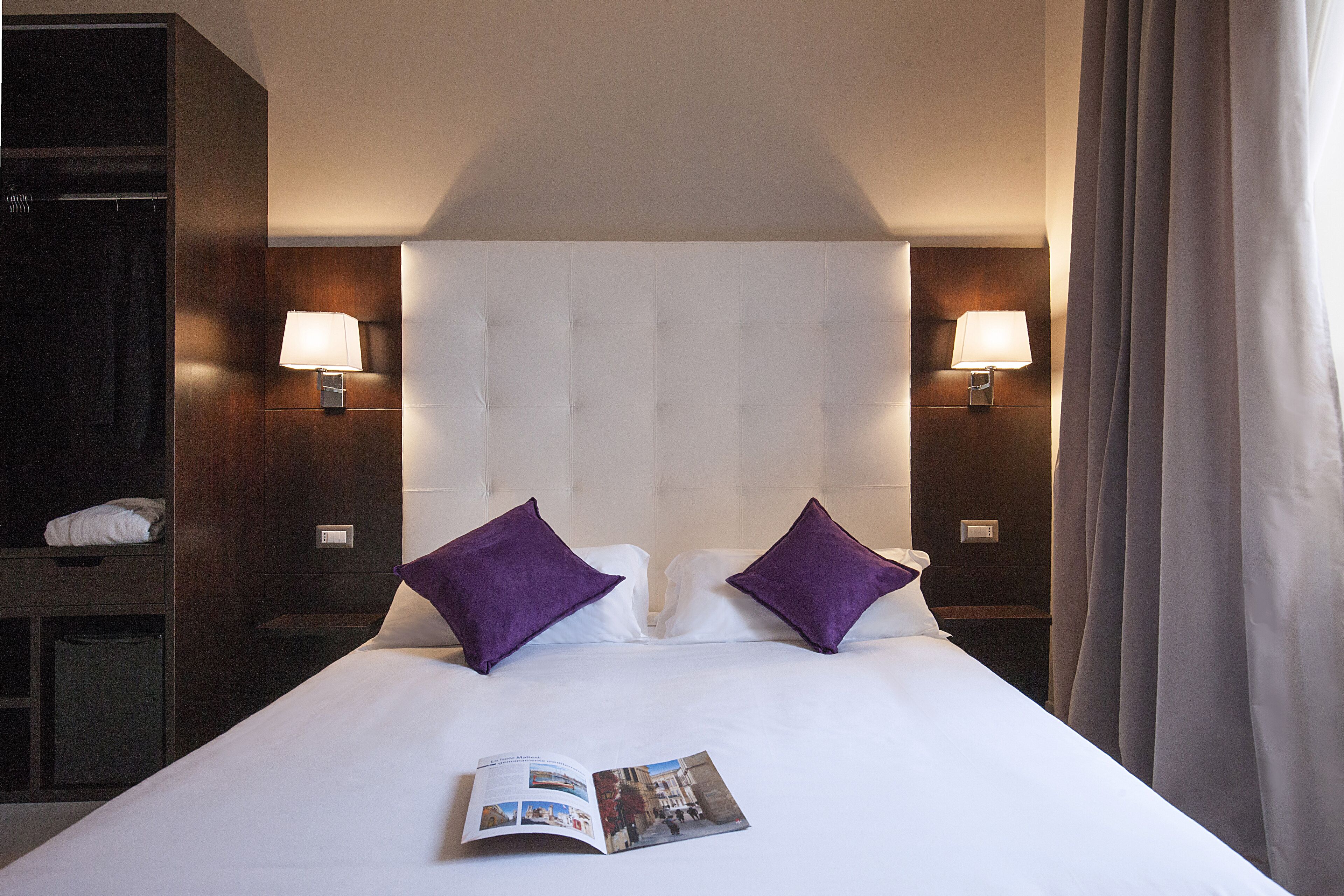 A hotel room with a large bed, two purple pillows, two bedside lamps, and a magazine on the bed.
