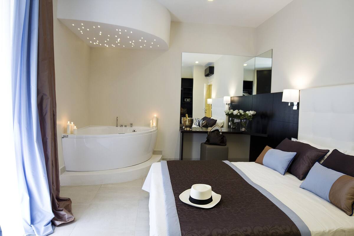 Habitación de hotel moderna con una cama grande, zona de estar separada, bañera y espejo.