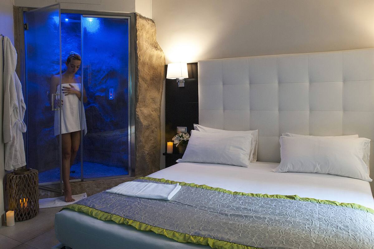 Una habitación de hotel con cama, mesita de noche, lámpara fijada a la pared y zona de ducha con paredes de vidrio.
