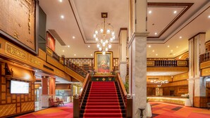 A grand hotel lobby with a red carpeted staircase, a large chandelier, and a prominent painting above the stairs. - Alexander Hotel (Bangkok)
