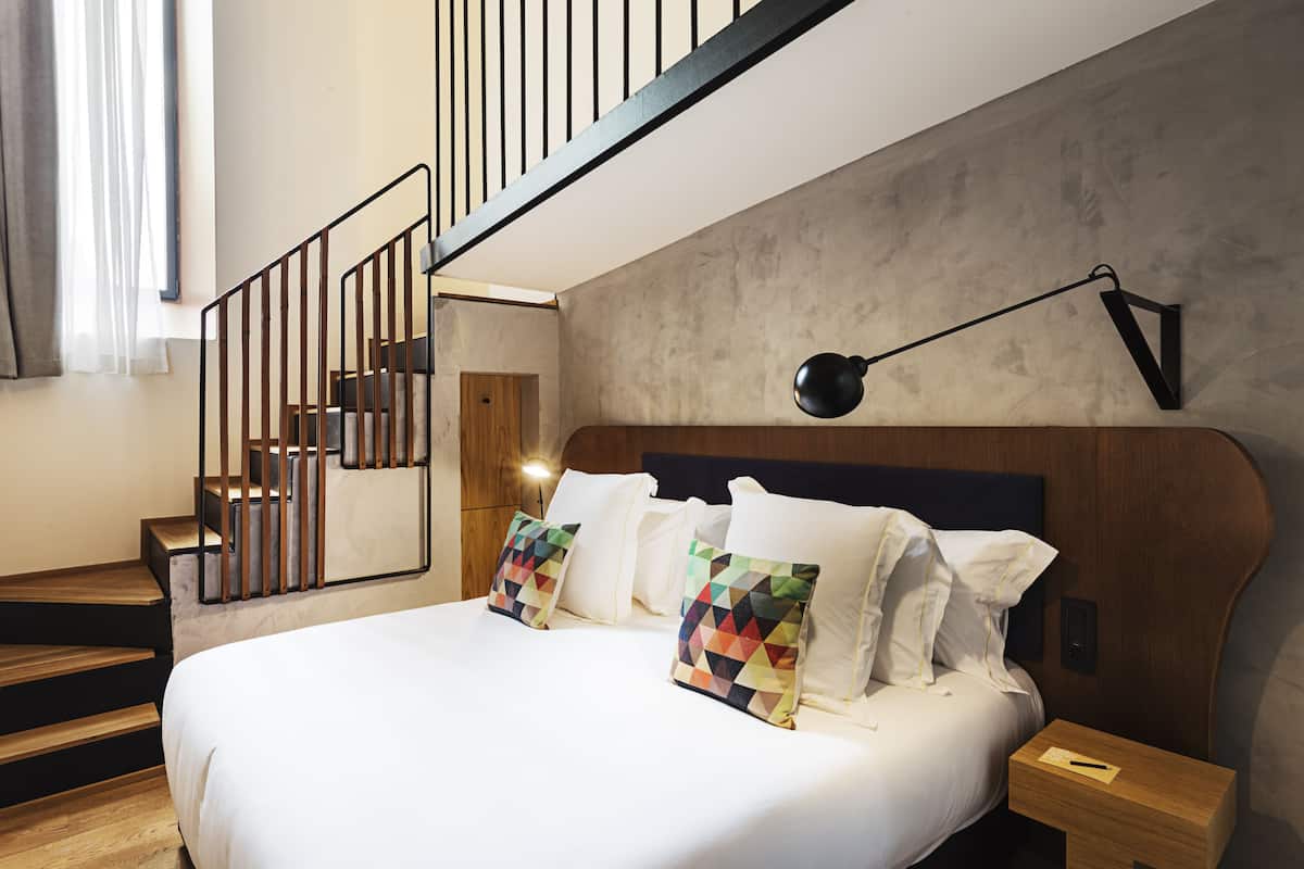 A modern hotel room with a large bed, wooden staircase, and a wall-mounted lamp.
