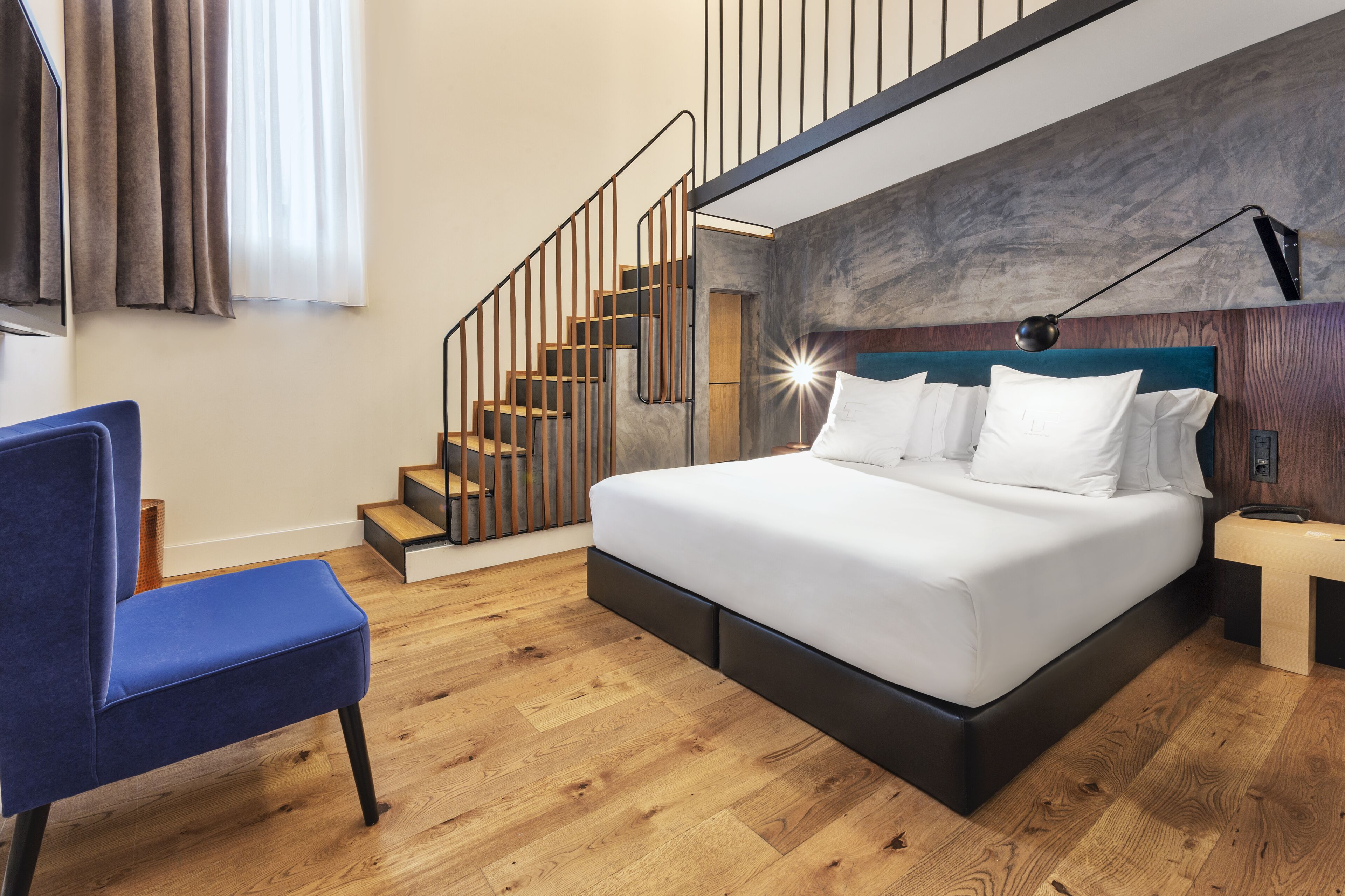 A modern bedroom with a large bed, a blue chair, a wooden staircase, and a nightstand.