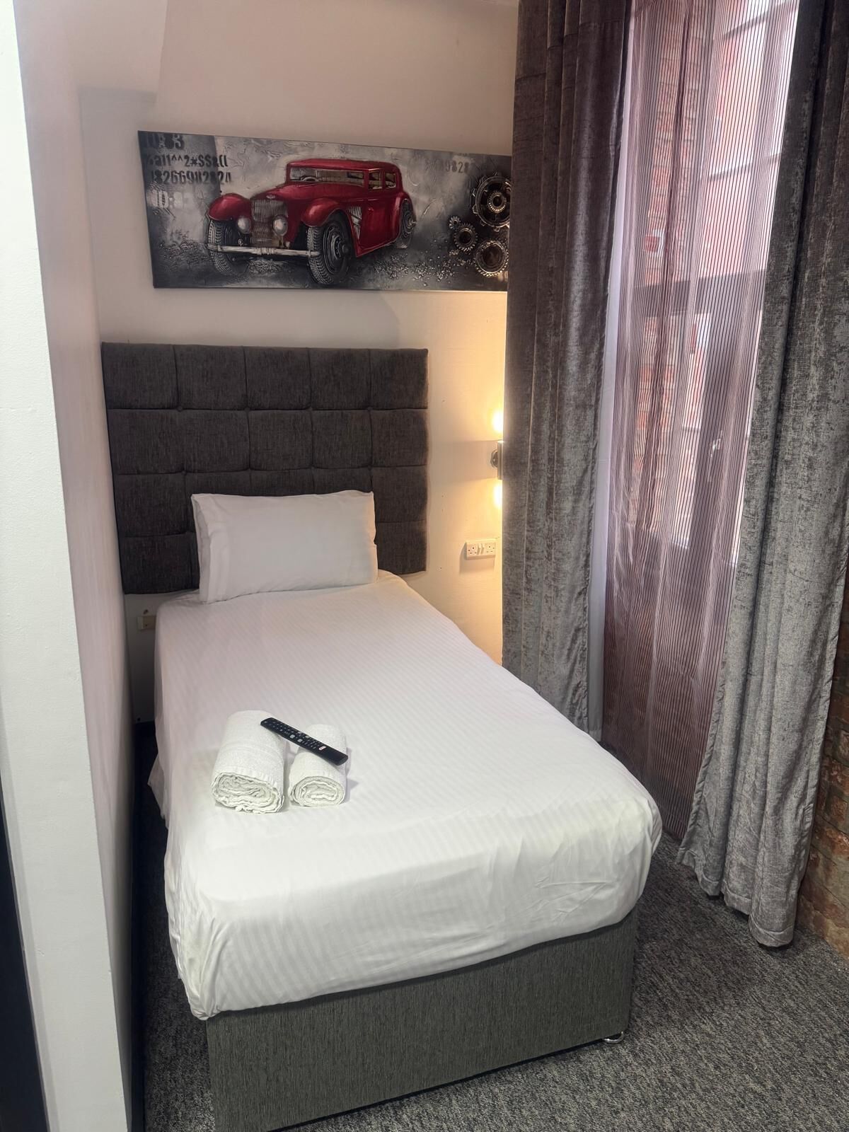A neatly made bed with a remote control and rolled towels, a grey upholstered headboard, and a wall-mounted artwork of a vintage car.