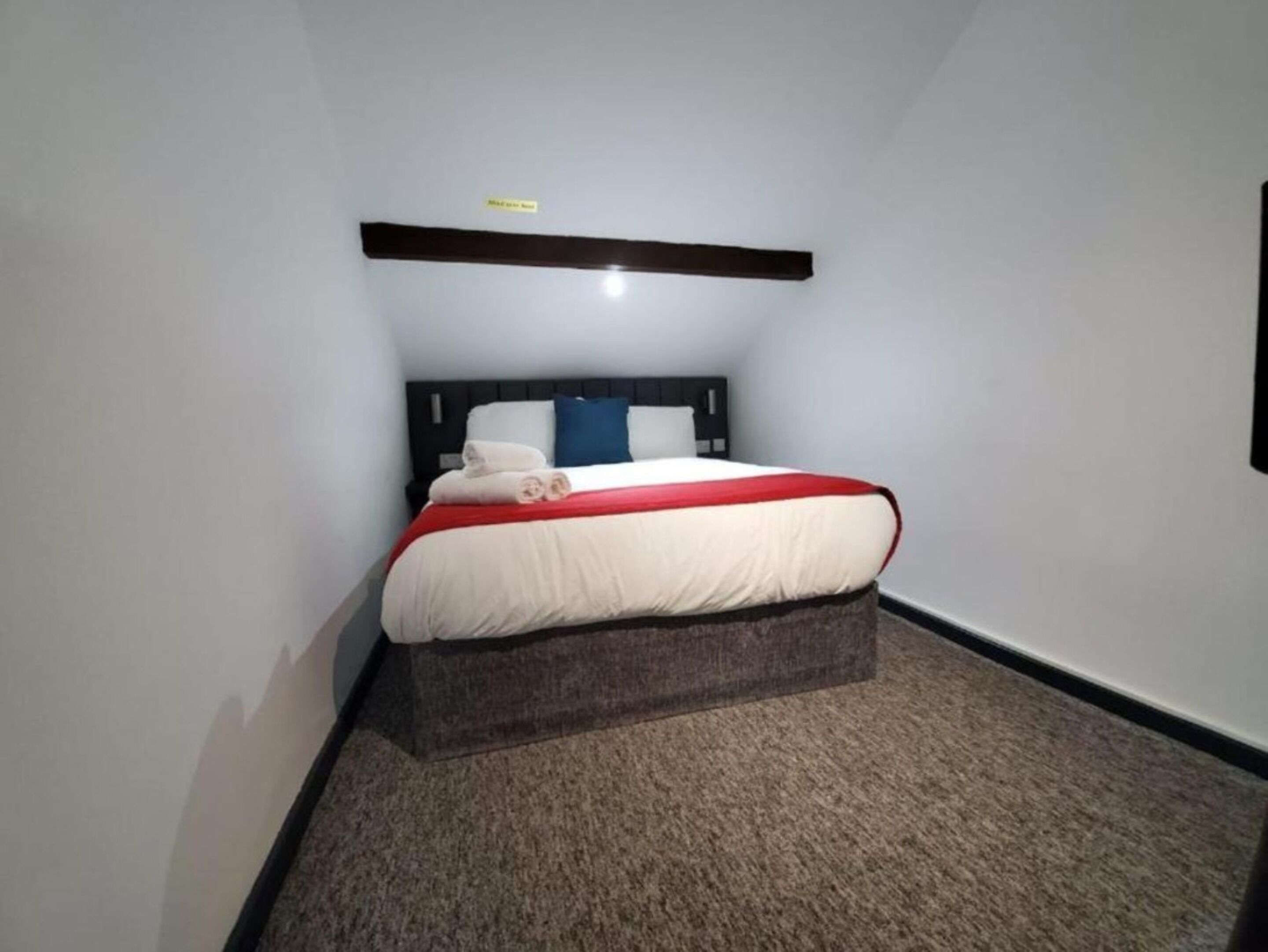 A bedroom with a bed, a headboard, a red and white bedspread, and a brown carpet.