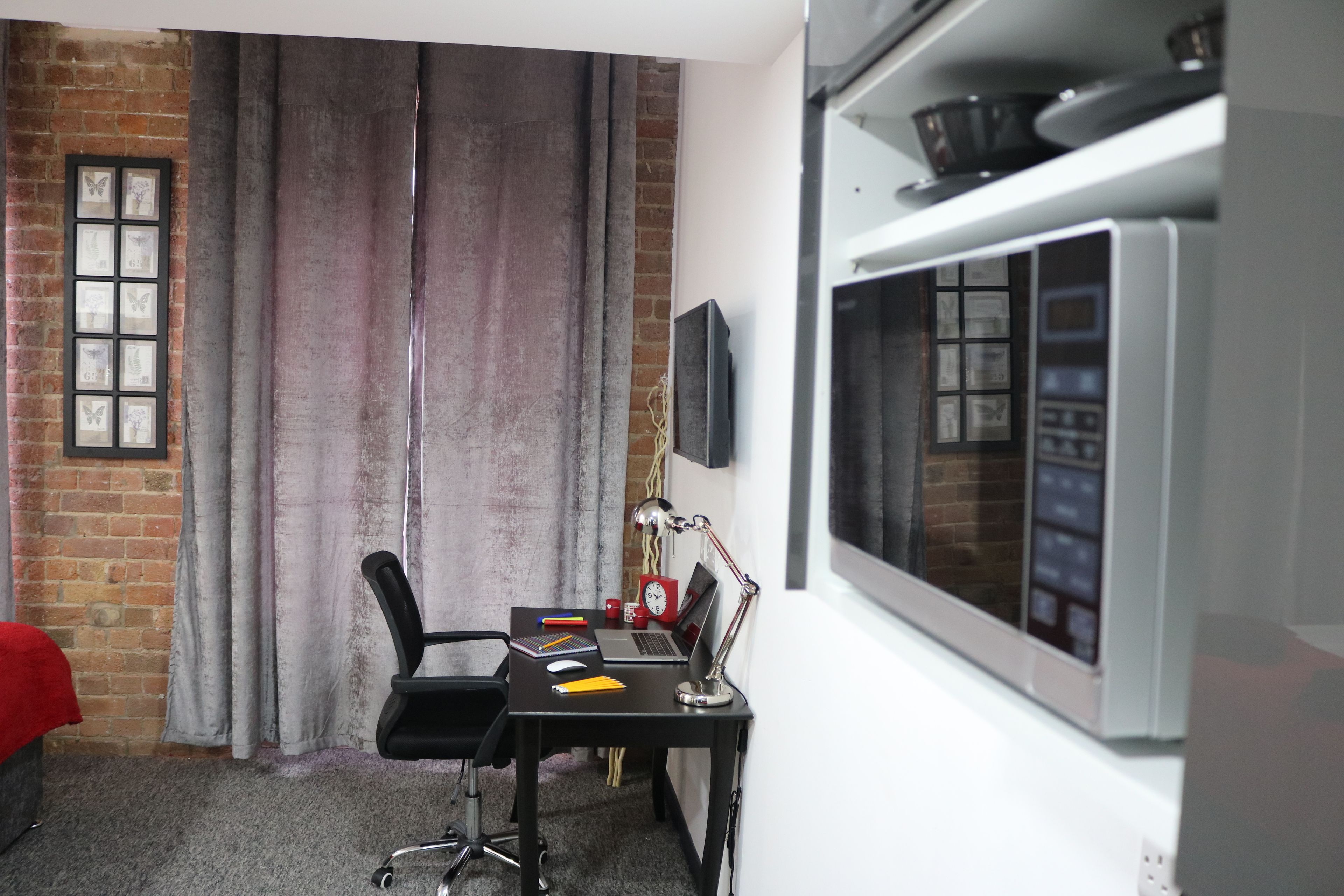A room with a brick wall, a black desk with a laptop, a red chair, a black office chair, a window with blinds, and a white shelf with a microwave.