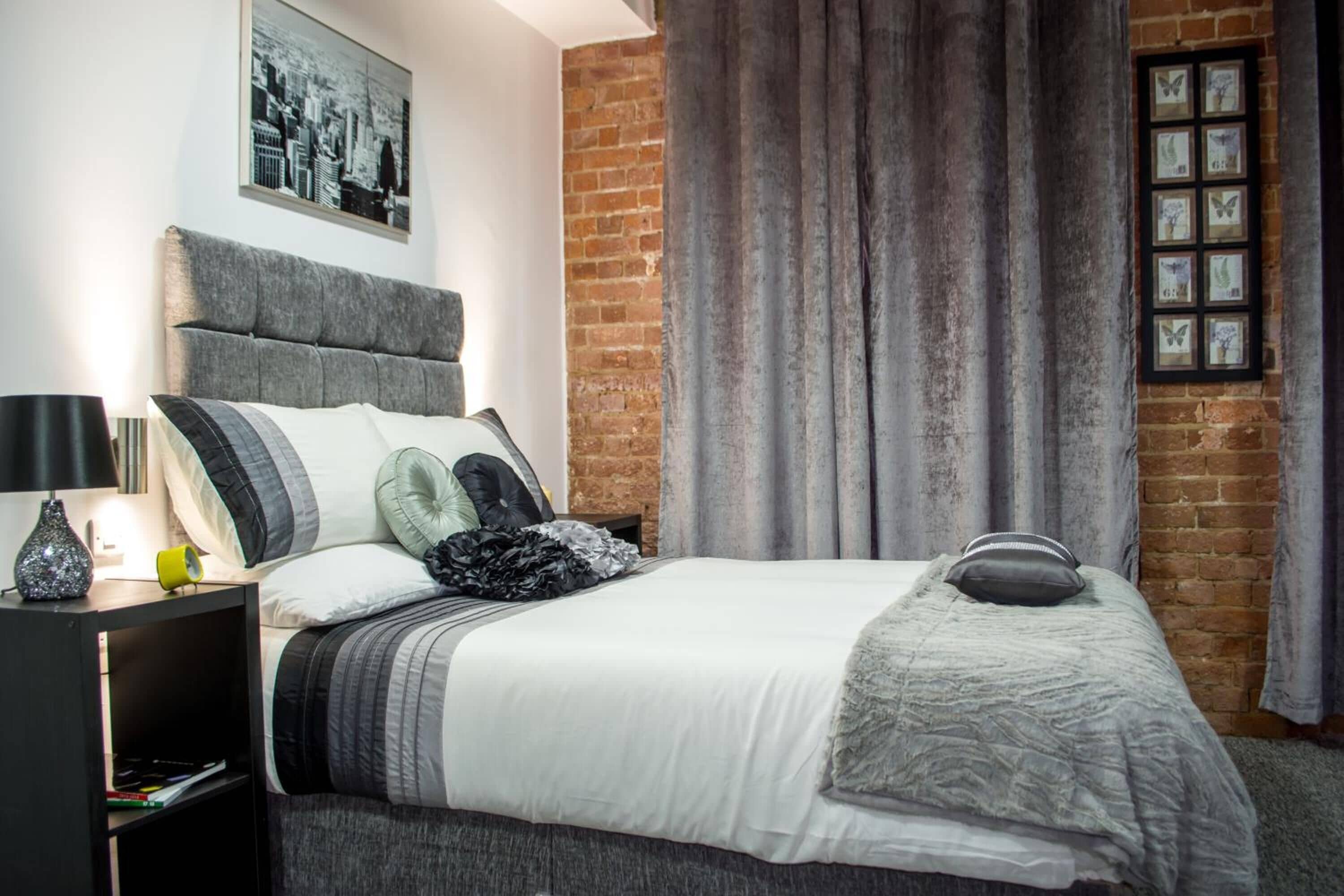 A modern bedroom with a brick wall, a large bed with grey bedding, a black nightstand with a lamp, and a framed cityscape picture above the bed.
