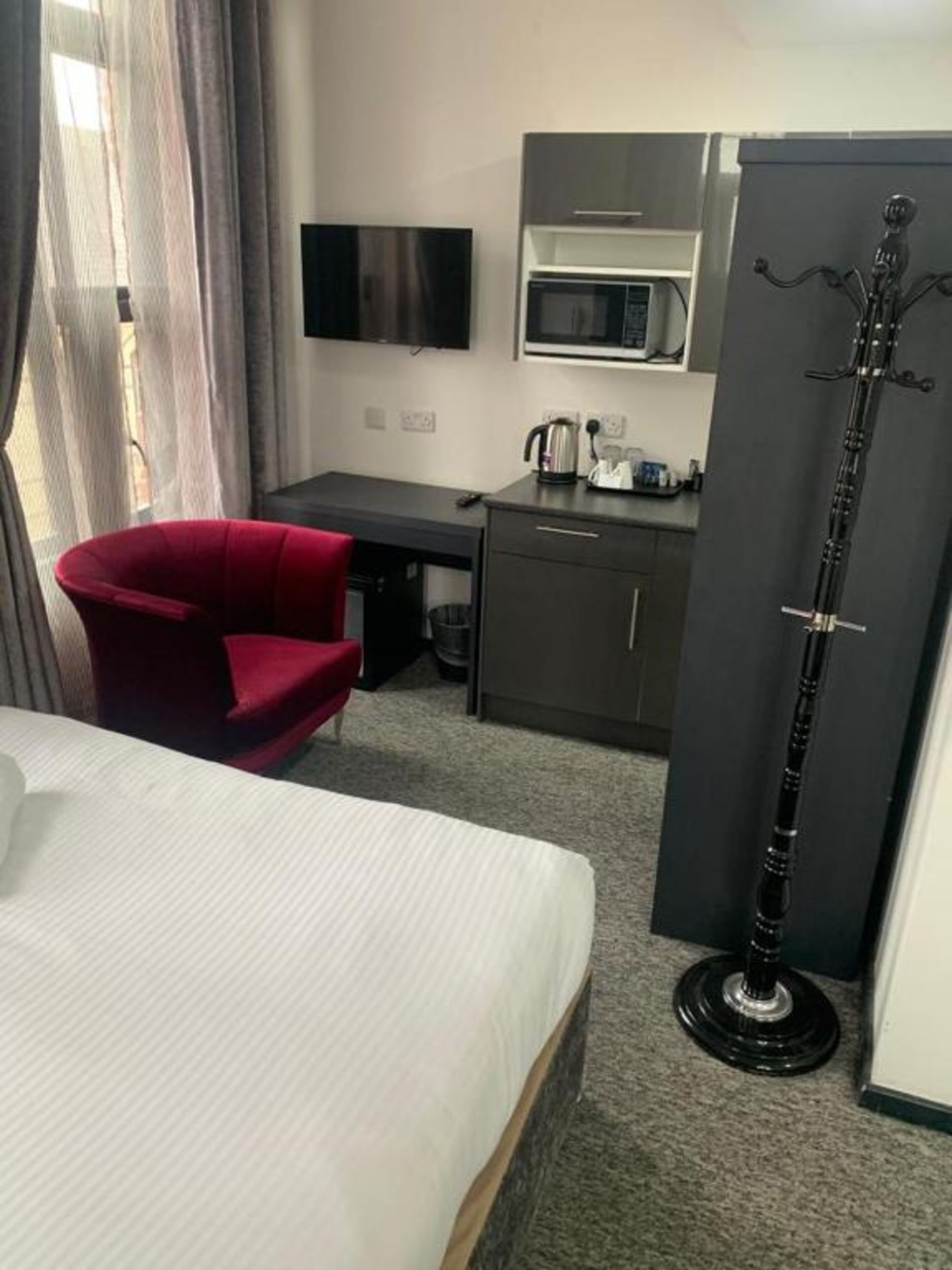 Een hotelkamer met een bed, een rode stoel, een bureau, een kitchenette en een hoge, zwarte vloerlamp.
