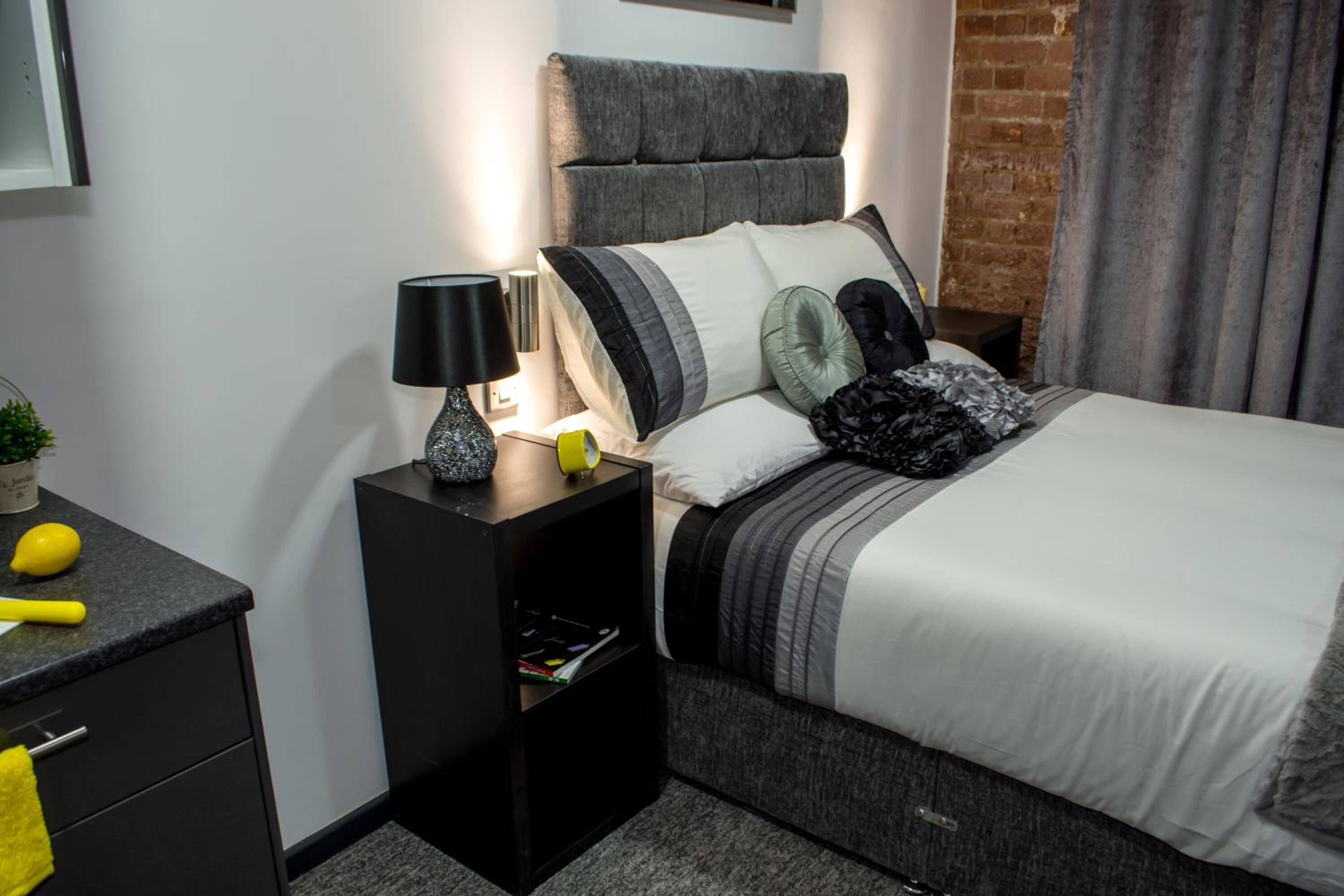 A modern bedroom with a grey upholstered headboard, a bedside table with a lamp, and a bed with striped bedding and decorative pillows.