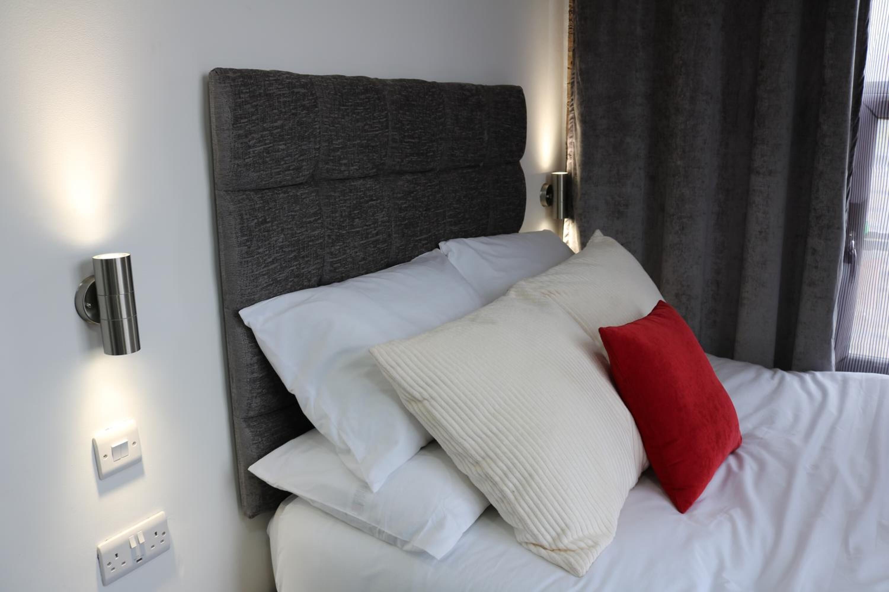 Ein ordentlich bezogenes Bett mit weißen und roten Kissen, einem grauen Kopfteil und einer Wandleuchte.