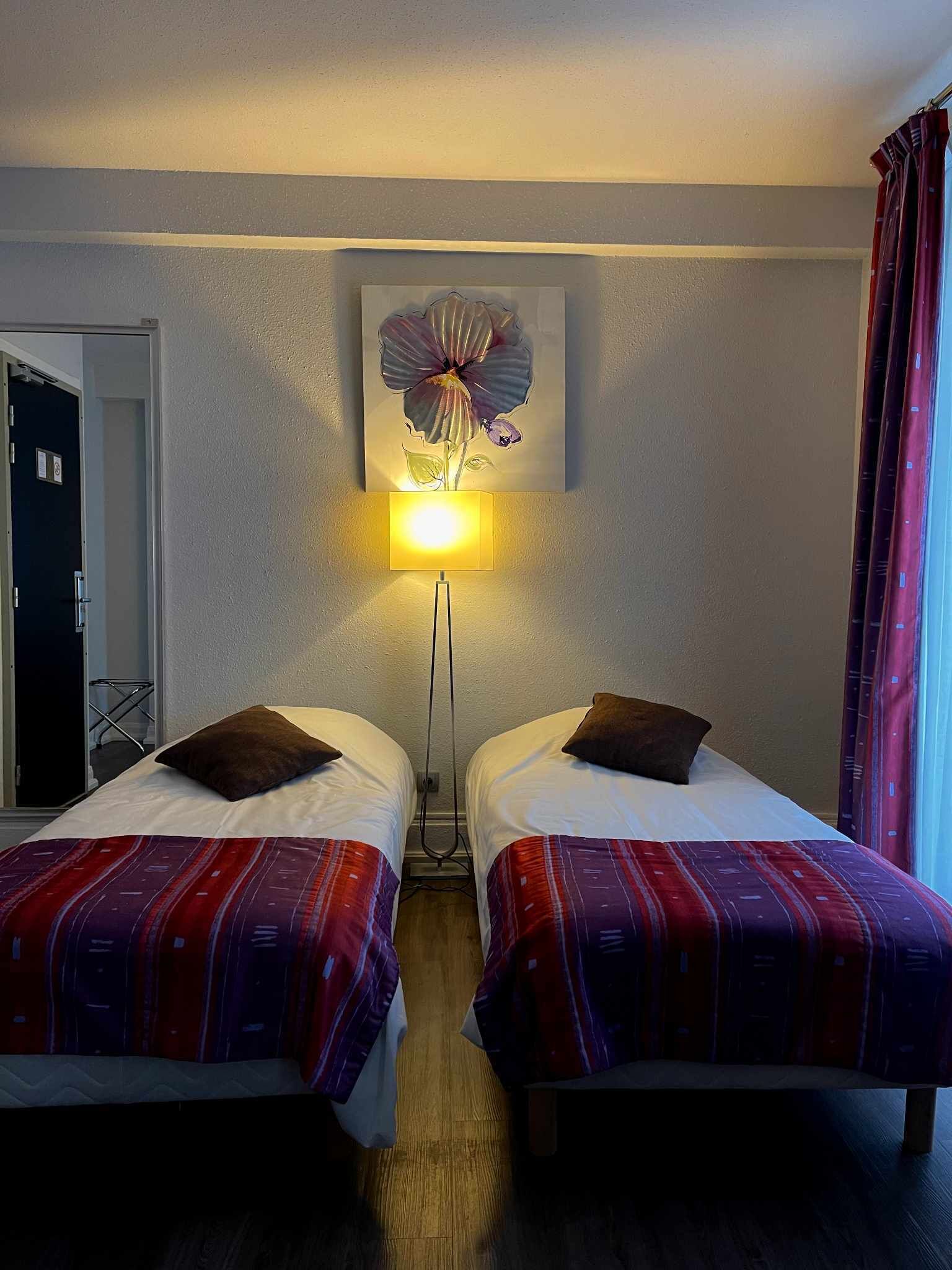 A hotel room with two beds, a wall-mounted lamp, and a door to the left.