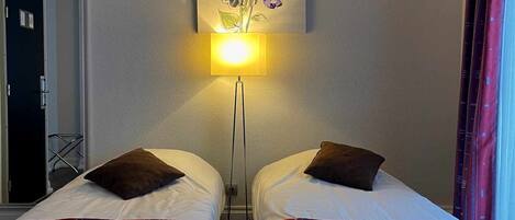 Habitación de hotel con dos camas, una lámpara en la pared y una puerta a la izquierda.