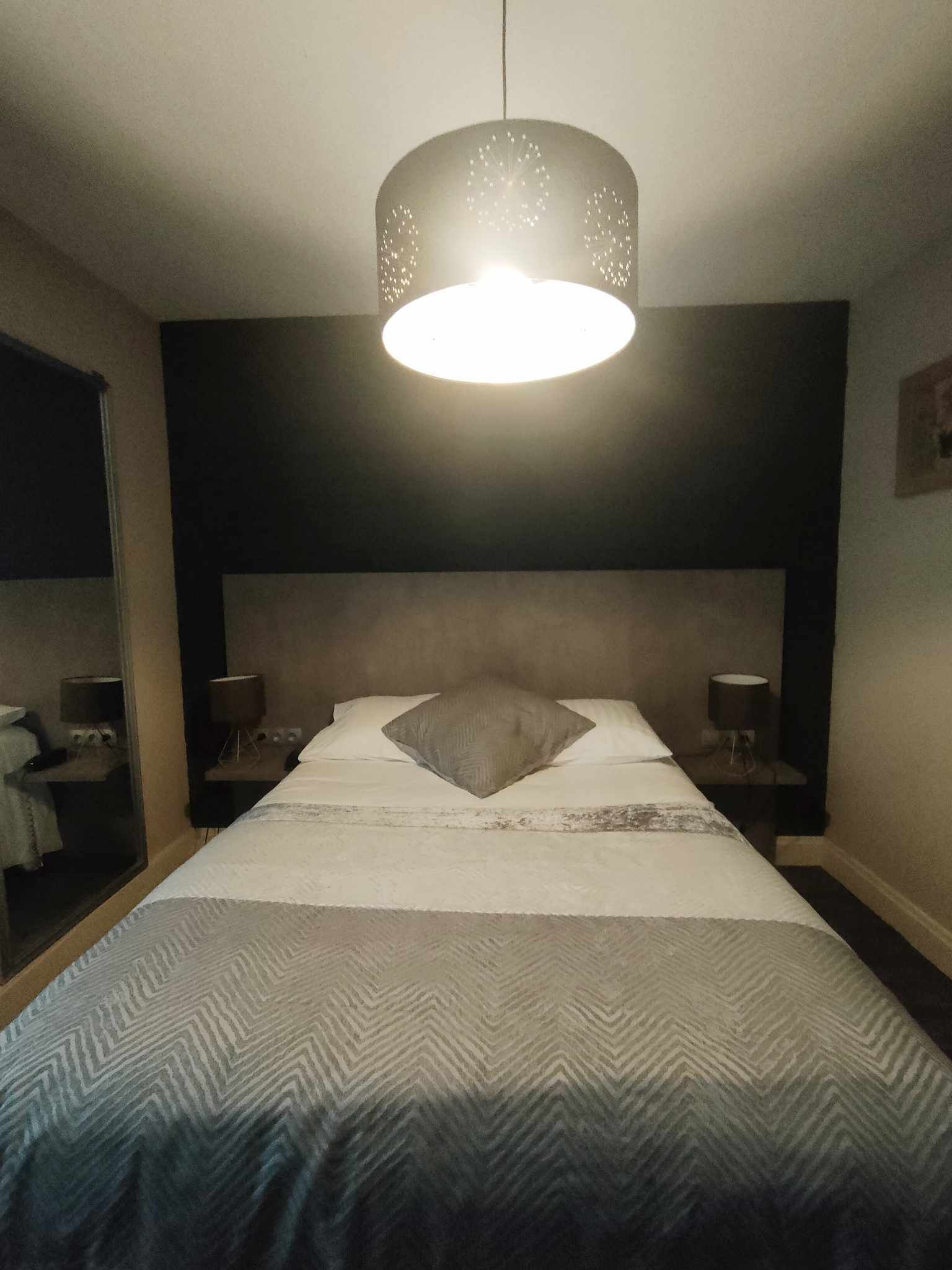 A neatly made bed with a patterned bedspread, a large round light fixture above, and a dark headboard.