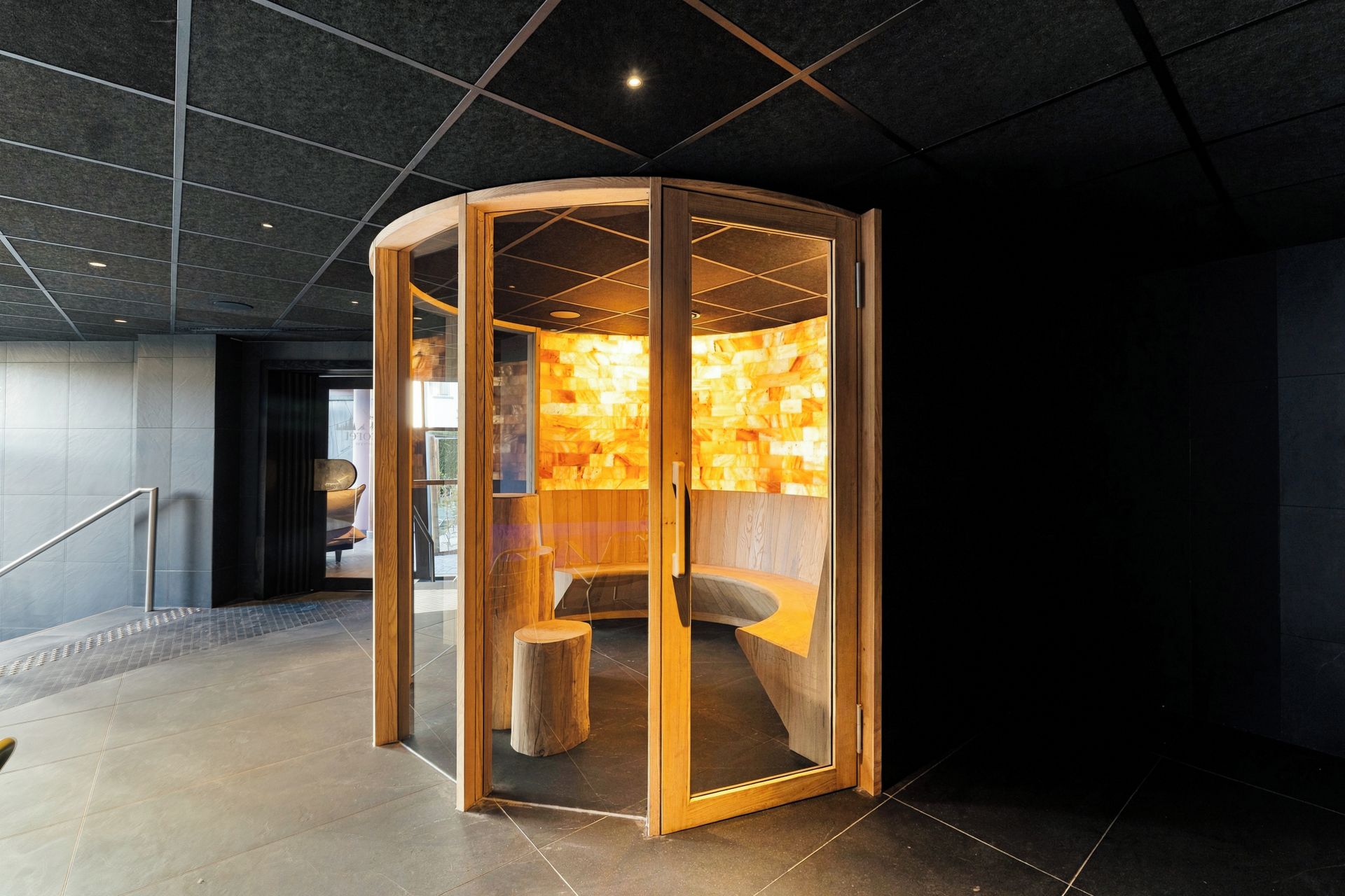 Un área de relajación moderna y cerrada con interior de madera y puerta de vidrio, ubicada en un vestíbulo amplio con piso de baldosas y una escalera.