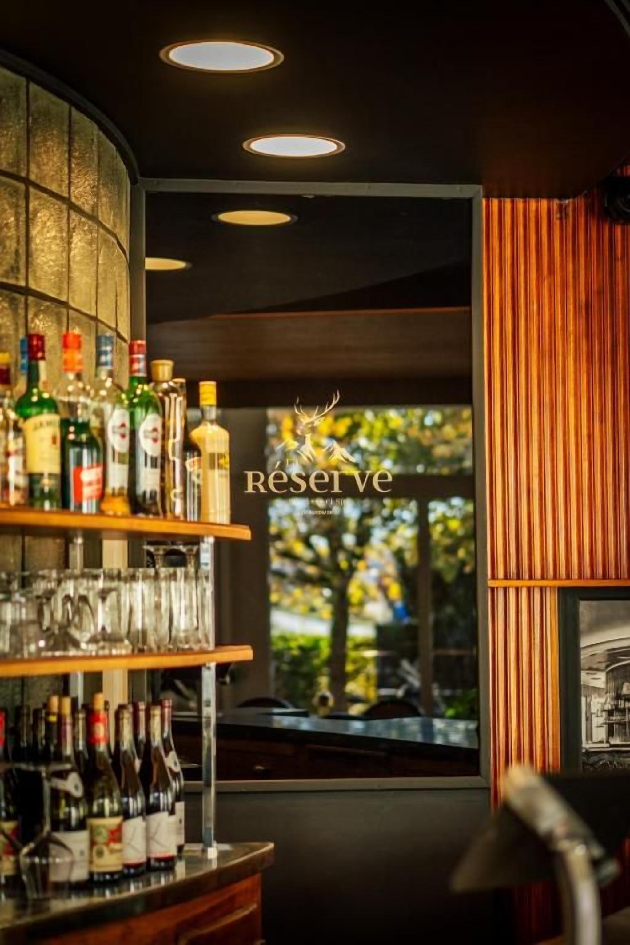 Una zona de bar con estantes llenos de botellas y vasos, un letrero que dice 'Reserve' y una vista por la ventana.