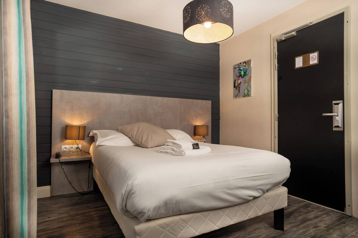 Una cama bien hecha con edredón blanco y una toalla doblada, una lámpara de noche y un aplique en la pared sobre la cama.