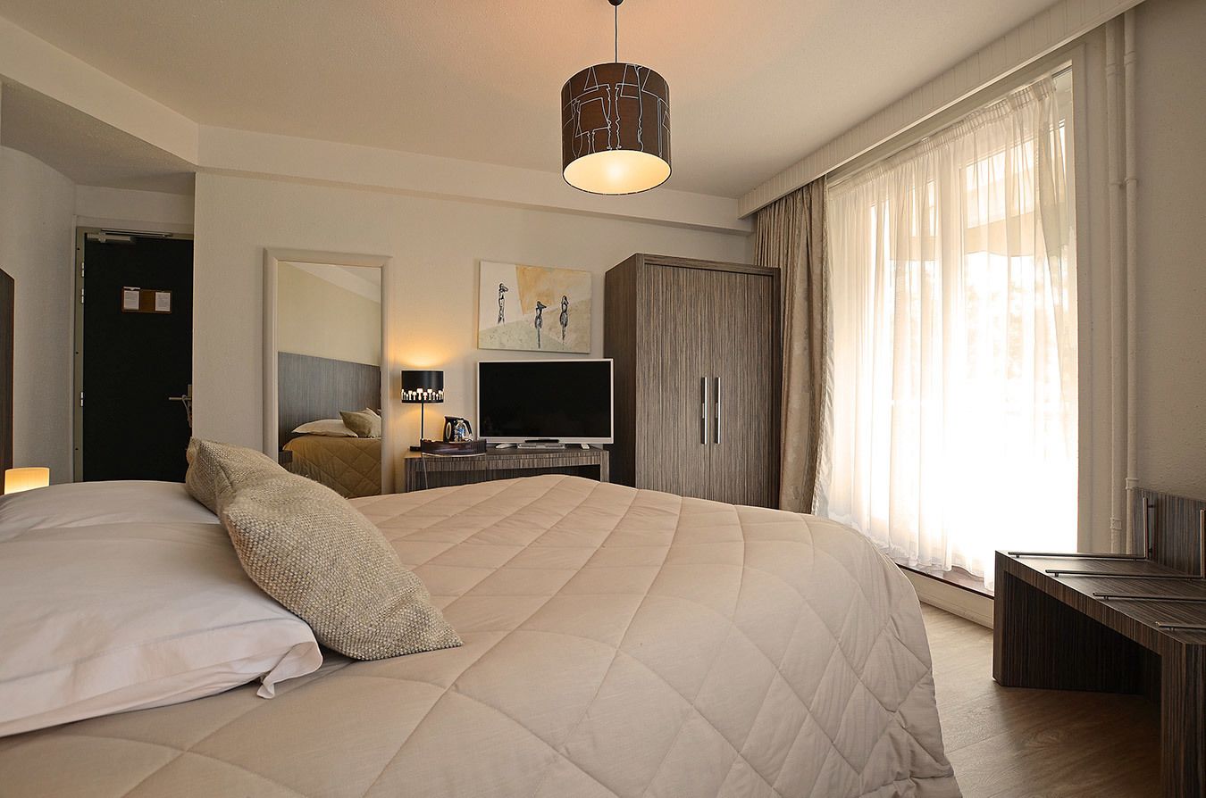 Habitación de hotel con una cama grande, un escritorio con televisor de pantalla plana, un armario y un ventanal con cortinas de gasa.