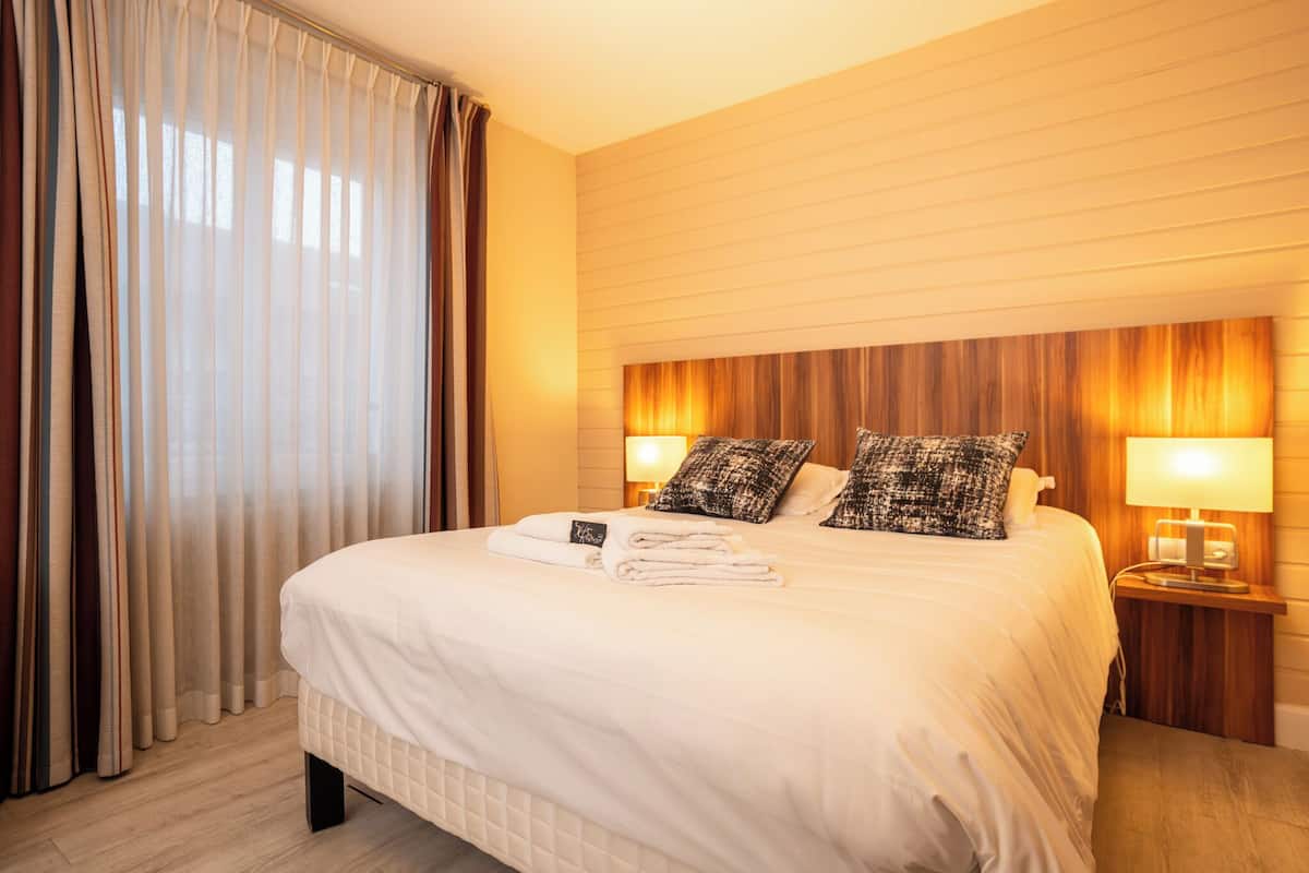Una habitación de hotel con una cama grande, dos lámparas de noche, un cabecero de madera y una ventana con cortinas.