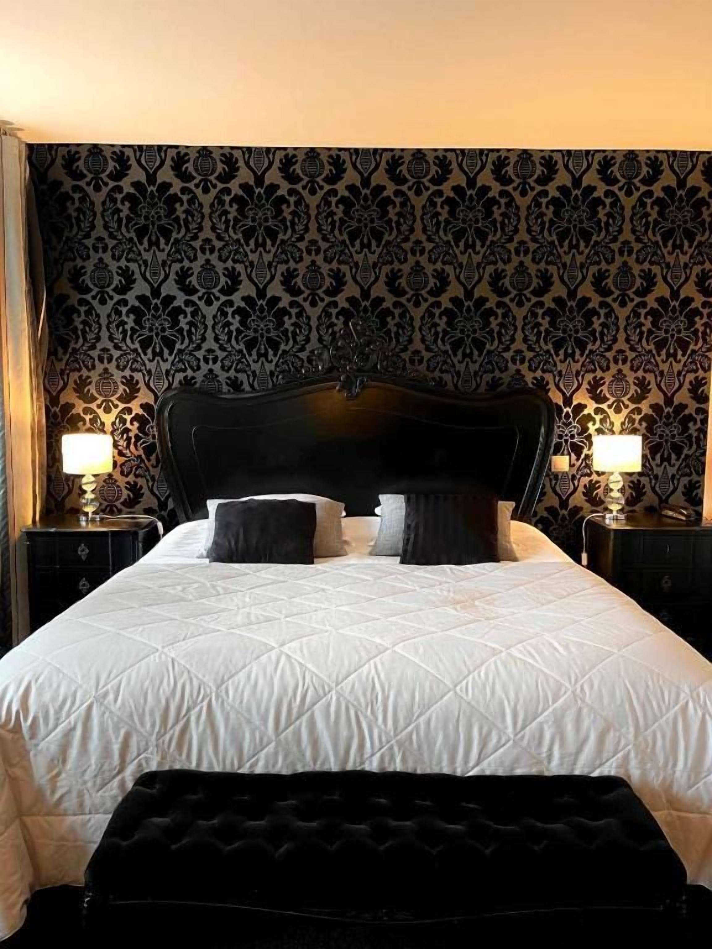 Ein Hotelzimmer mit einem großen Bett, zwei Nachttischlampen, einem Kopfteil und einem Wandpapier mit Muster.