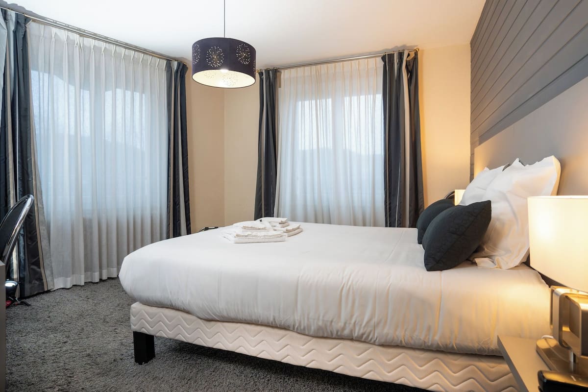 Habitación de hotel con una cama grande, una mesita de noche con lámpara y una ventana con cortinas.