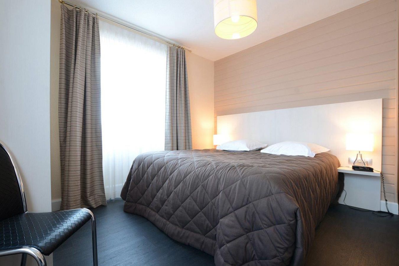 Een hotelkamer met een groot bed, een stoel, een nachtkastje met een lamp, en een raam met gordijnen.