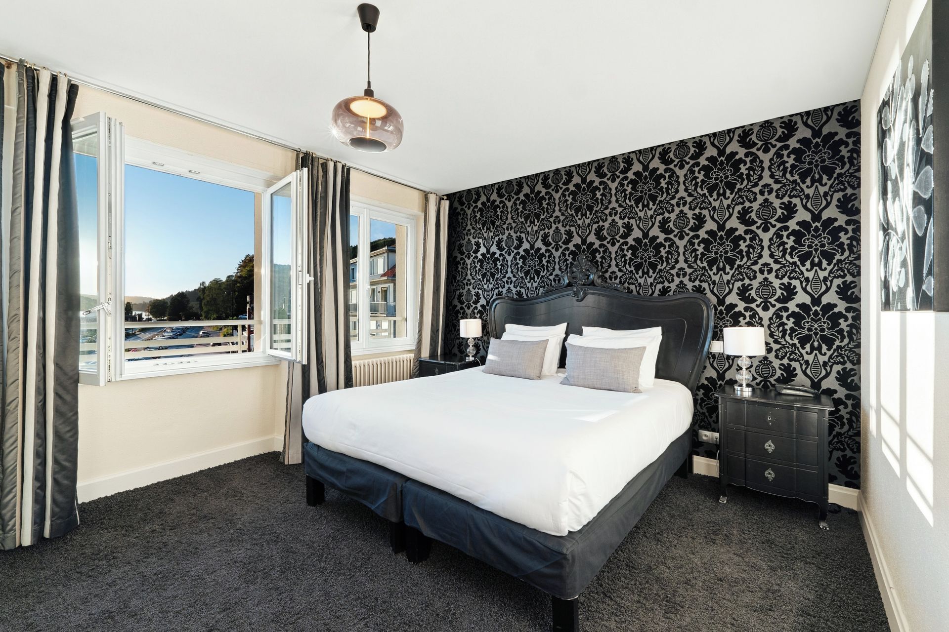 A hotel room with a large bed, a window with curtains, a patterned wall, and a nightstand with a lamp.