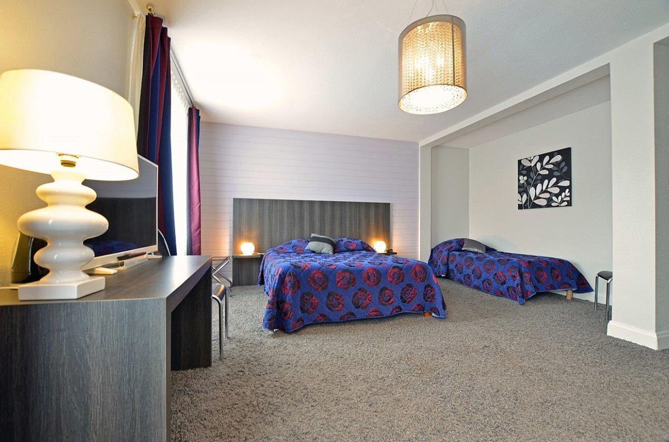 Hotelkamer met twee bedden, een bureau met lamp, een groot raam met gordijnen, een wandtelevisie en een plafondlamp.