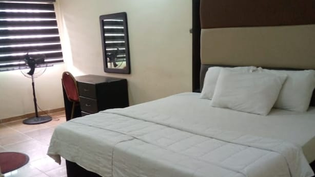 Una cama bien hecha con ropa blanca, un cabecero oscuro y un pequeño escritorio con una silla a un lado.