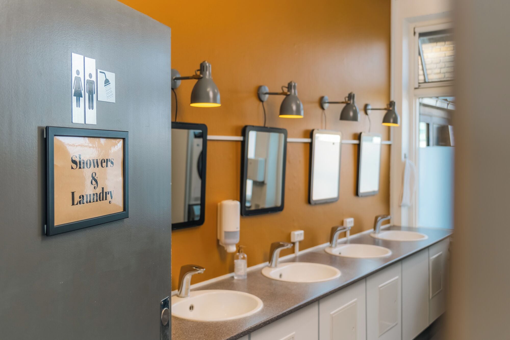 Une salle de bain moderne avec trois lavabos, des miroirs et un éclairage au plafond. Un panneau indique la présence de douches et d’installations de blanchisserie.