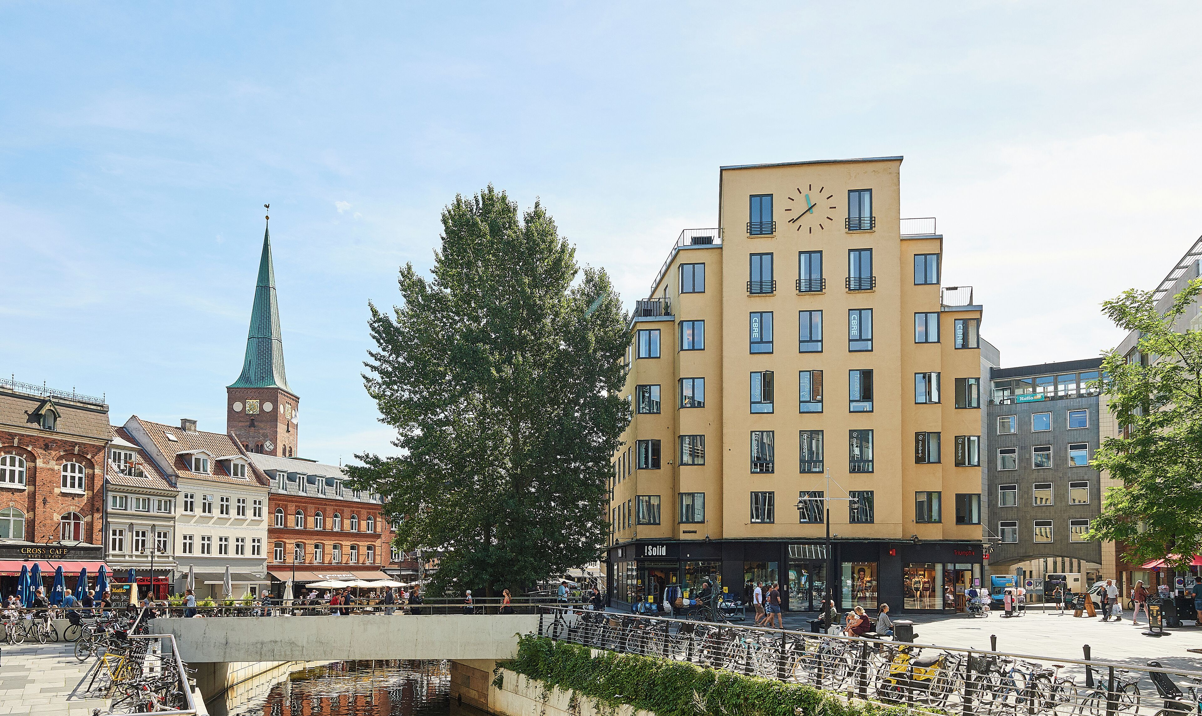 Une place de ville avec un pont, le clocher d’une église et des bâtiments modernes.