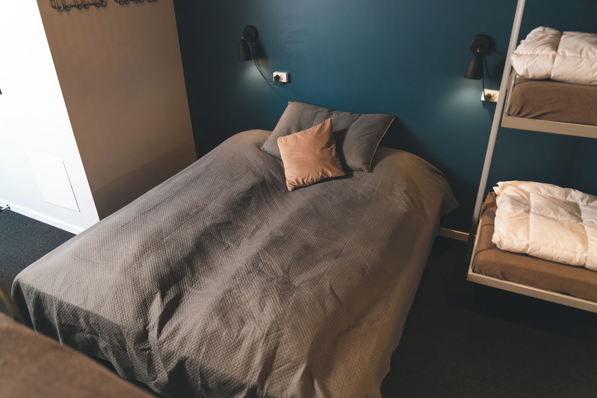 Una cama individual con una colcha texturizada y una almohada, una lámpara de noche y un estante en la pared con mantas dobladas.